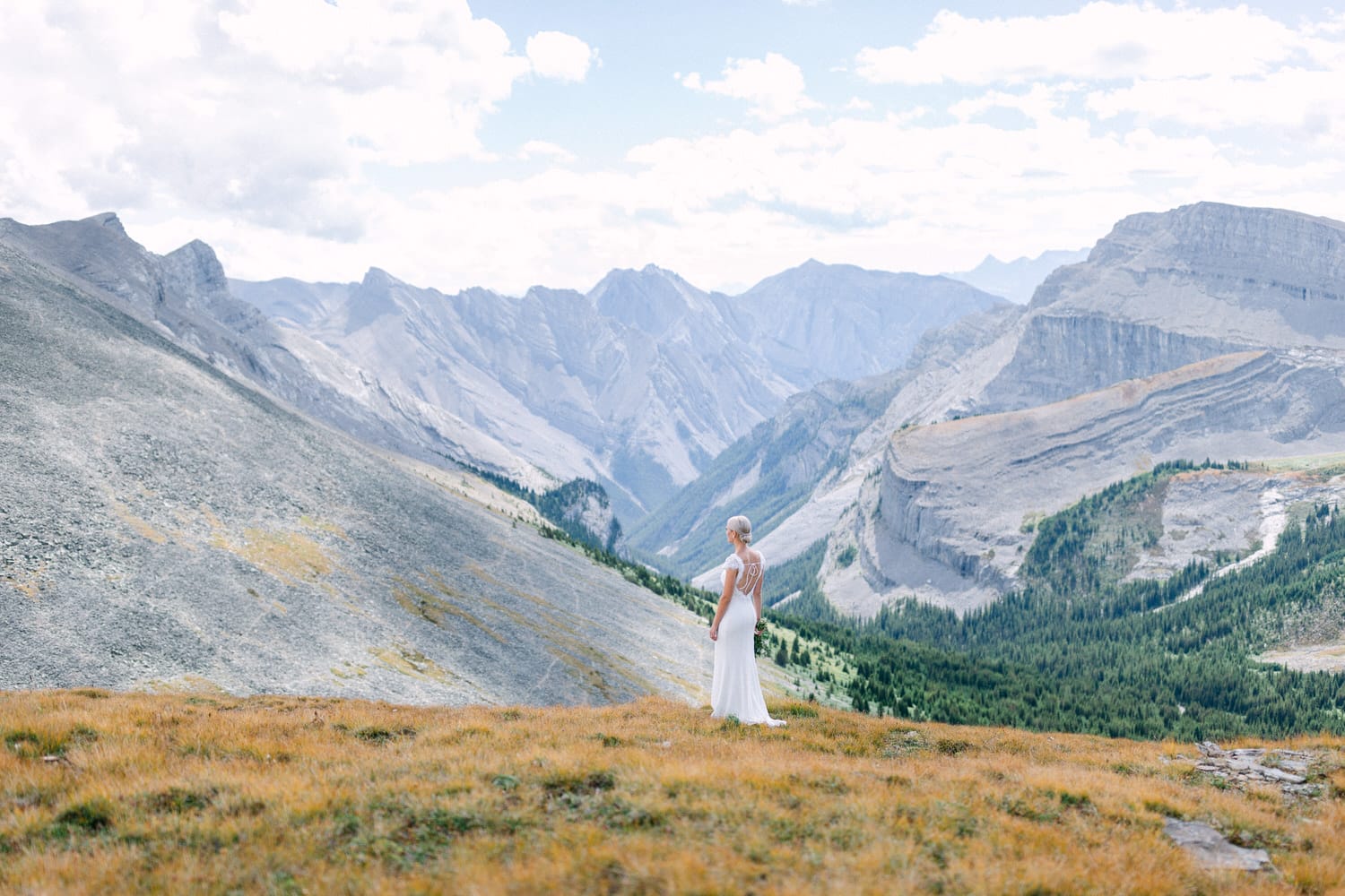 A bride in a flowing white gown stands on a hillside, overlooking a stunning valley surrounded by towering mountains and dramatic skies.