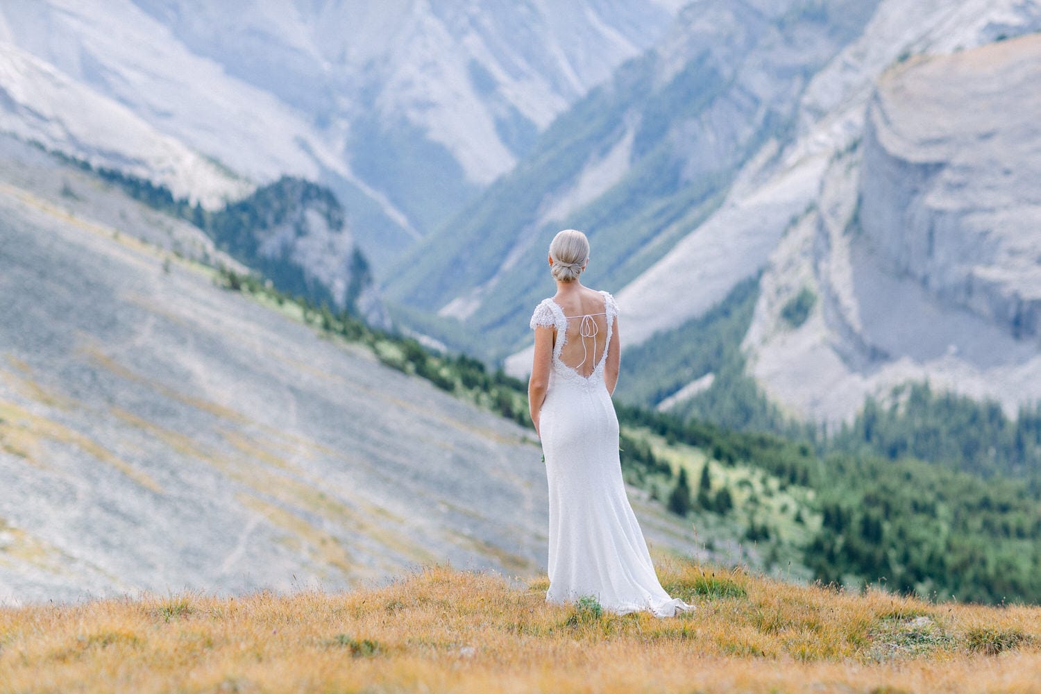 A bride in a white gown standing on a mountain overlook, facing away, with lush green hills and rocky peaks in the background.