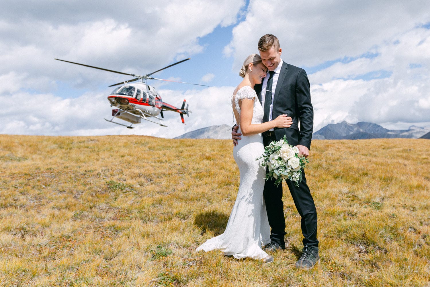 A bride and groom embrace in a scenic mountain landscape, with a helicopter hovering in the background.