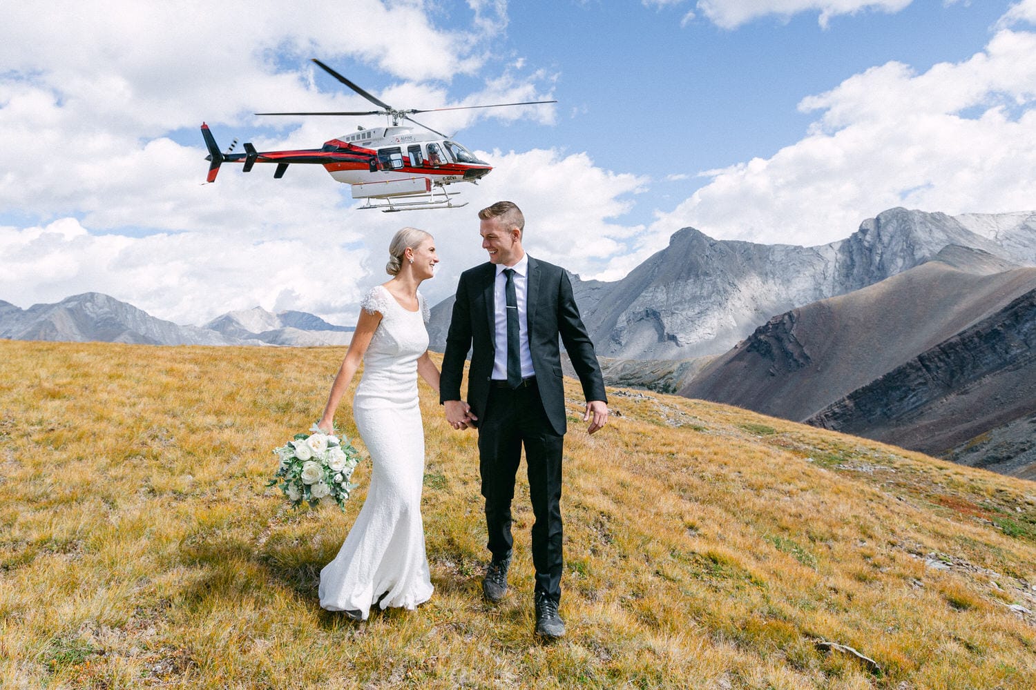 A joyful couple in wedding attire holding hands, surrounded by a picturesque mountain landscape, with a helicopter flying above.