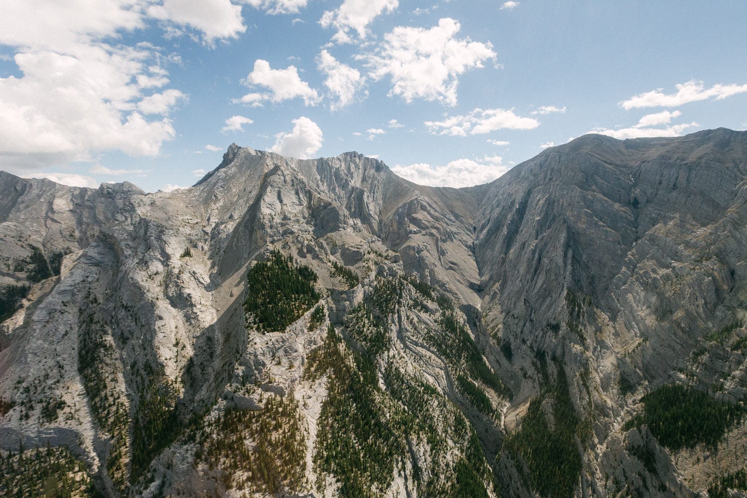 Aerial view of rugged mountains with green vegetation and scattered clouds in a bright blue sky.