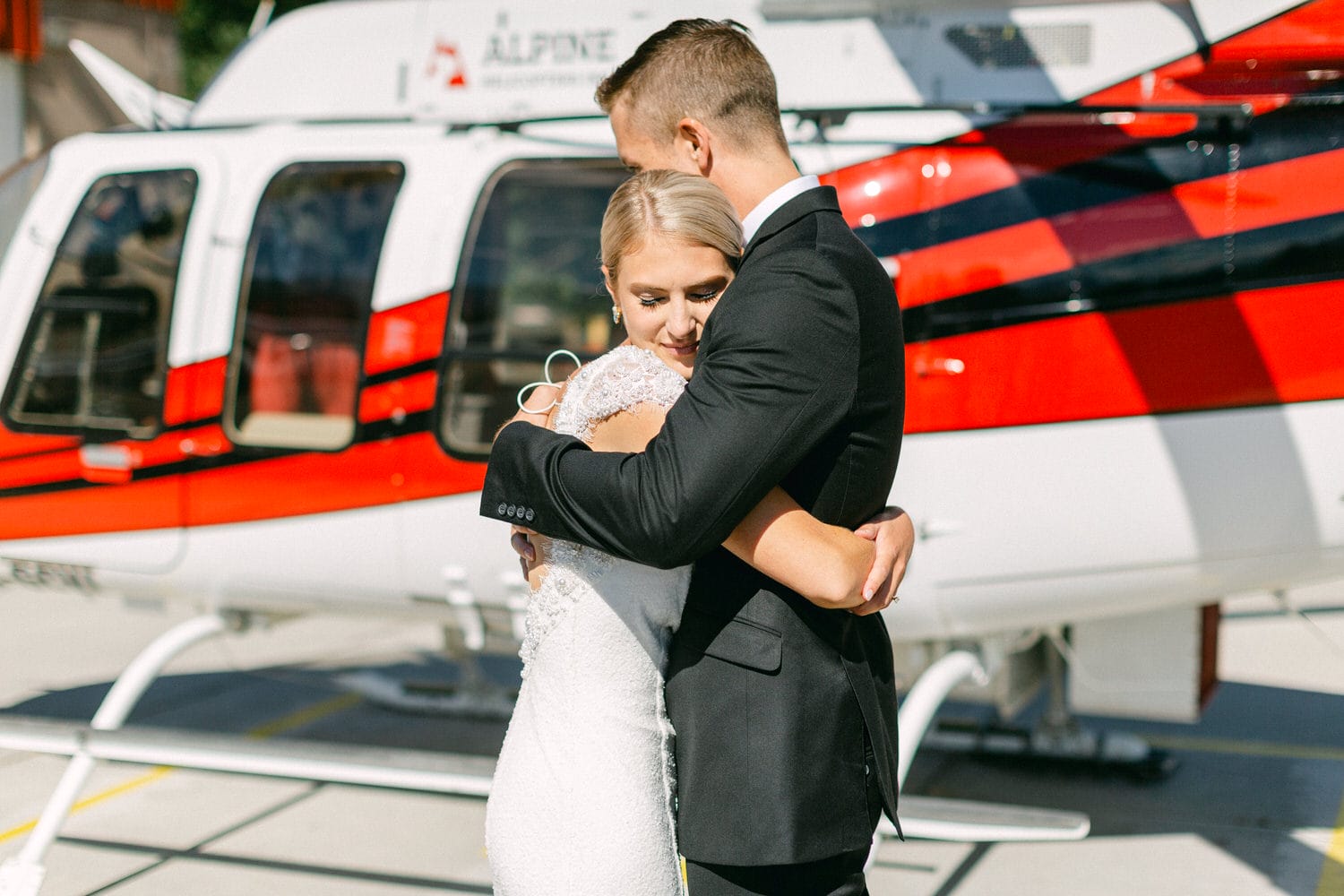 A bride and groom share a tender embrace in front of a vibrant red and white helicopter on a sunny day.