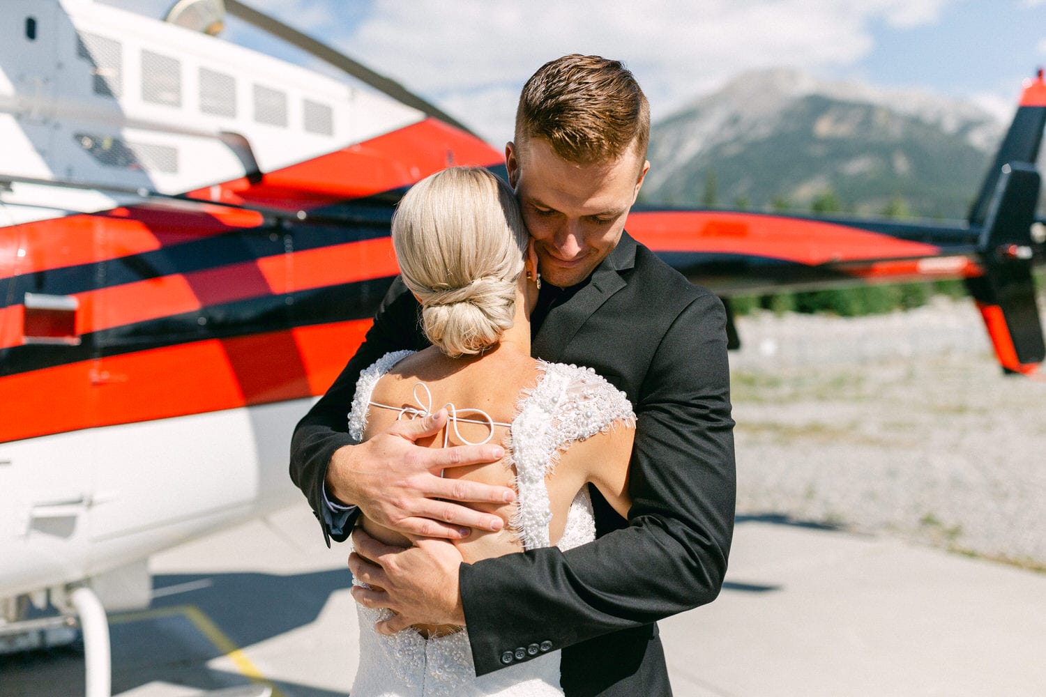 A couple shares a heartfelt embrace in front of a helicopter on a sunny day, showcasing love and connection against a beautiful mountain backdrop.