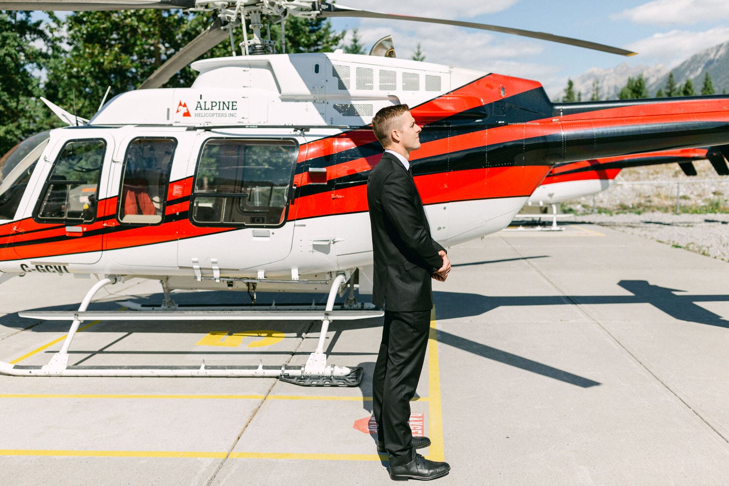 A man in a suit stands beside a red and white helicopter, preparing for departure on a sunny day.