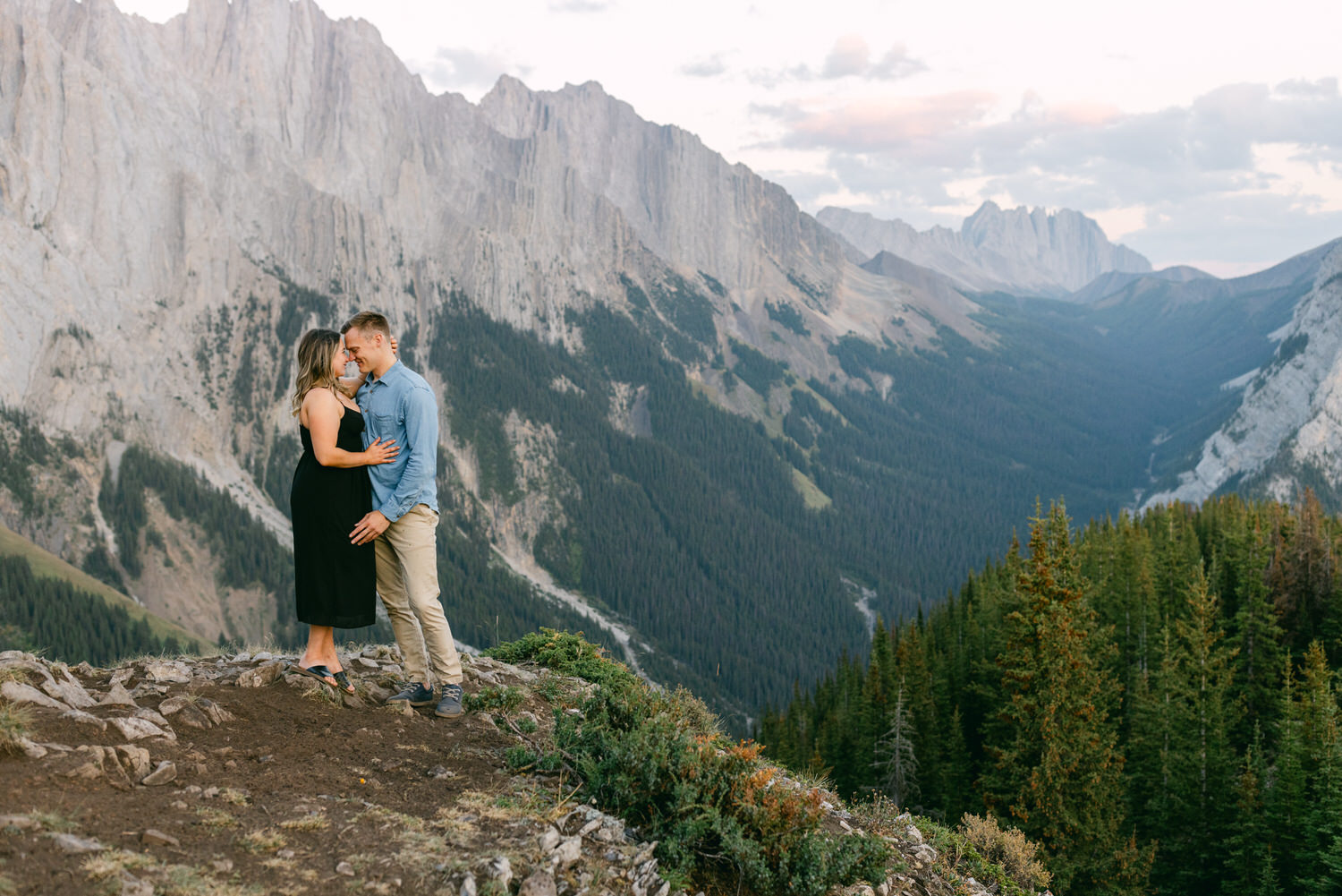 A couple shares an intimate moment on a rocky outcrop surrounded by breathtaking mountain scenery, showcasing their connection against a backdrop of lush forests and towering peaks.