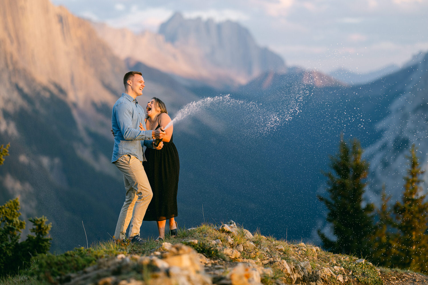 A joyful couple sprays champagne while laughing on a mountain top, with stunning peaks and lush greenery in the background.