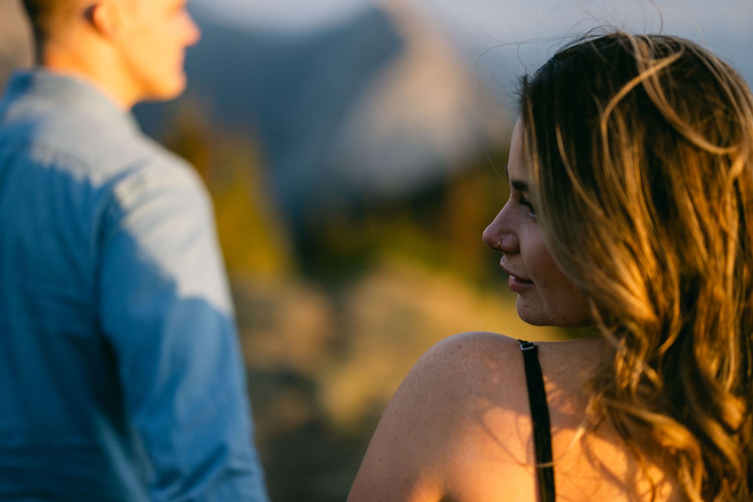 A woman gazes back at a man, with a scenic backdrop of mountains blurred in the distance, capturing a moment of connection in natural light.