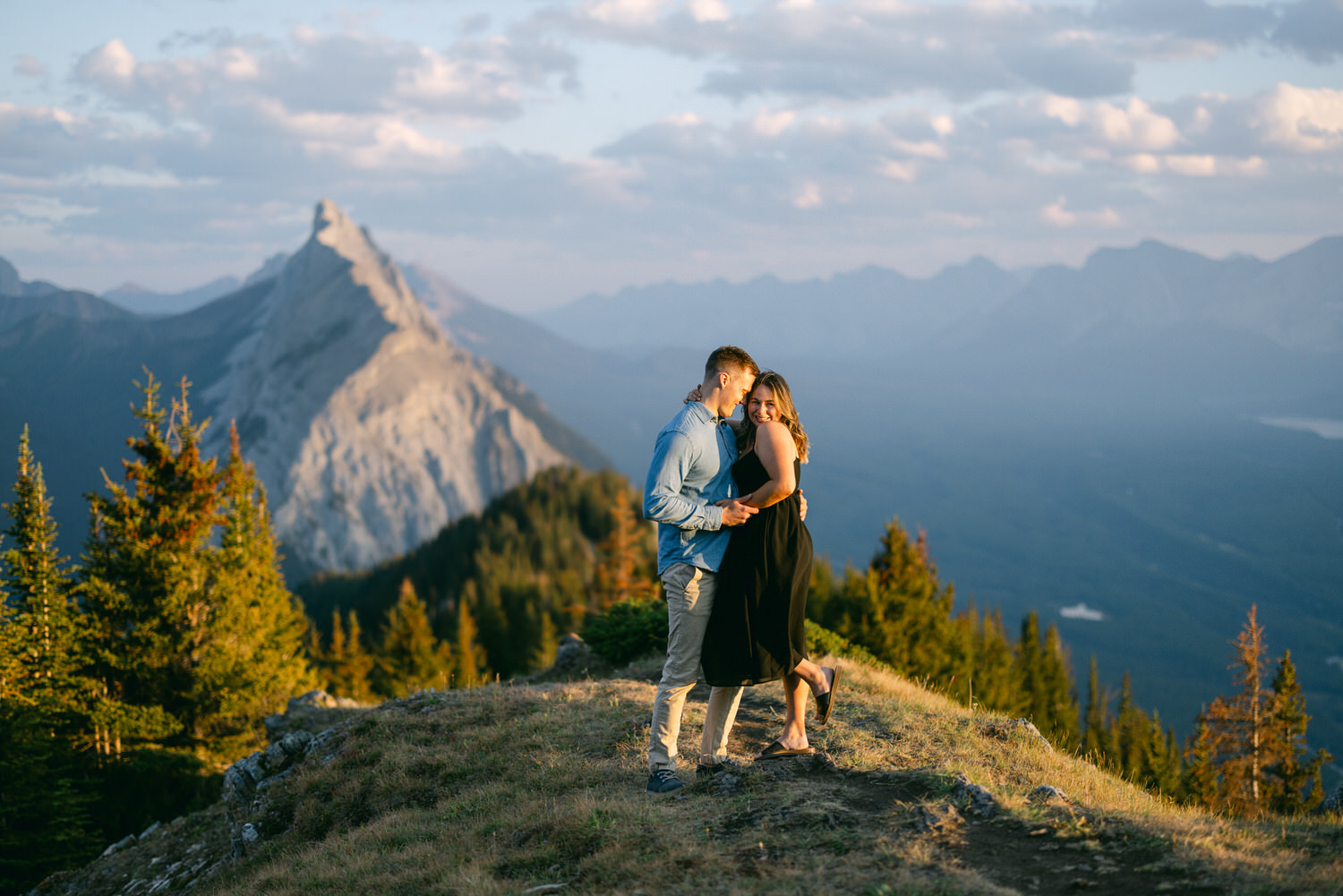 A couple stands together on a mountain ridge, surrounded by trees and mountains in the background, with a beautiful sunset illuminating the scene.