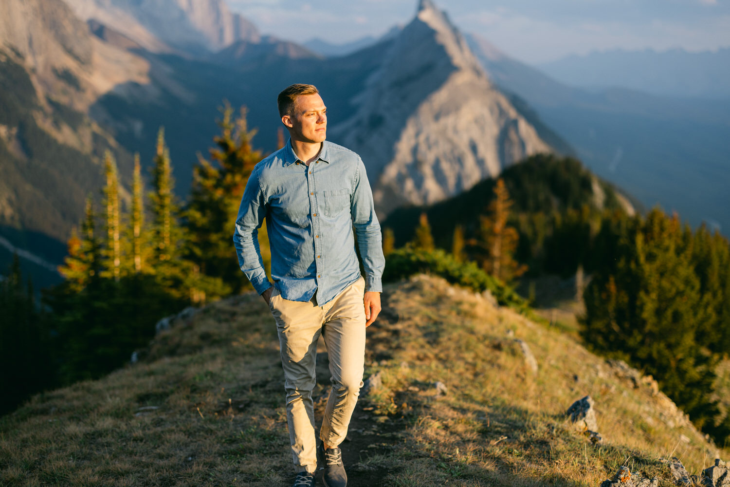 A man walks along a mountain ridge, surrounded by lush trees and majestic peaks under a clear sky.