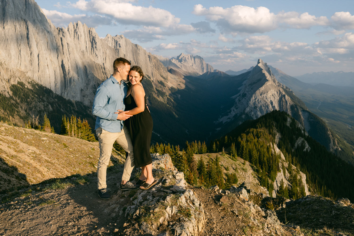 A couple shares a joyful moment while embracing on a rocky ledge with breathtaking mountains in the background under a partly cloudy sky.