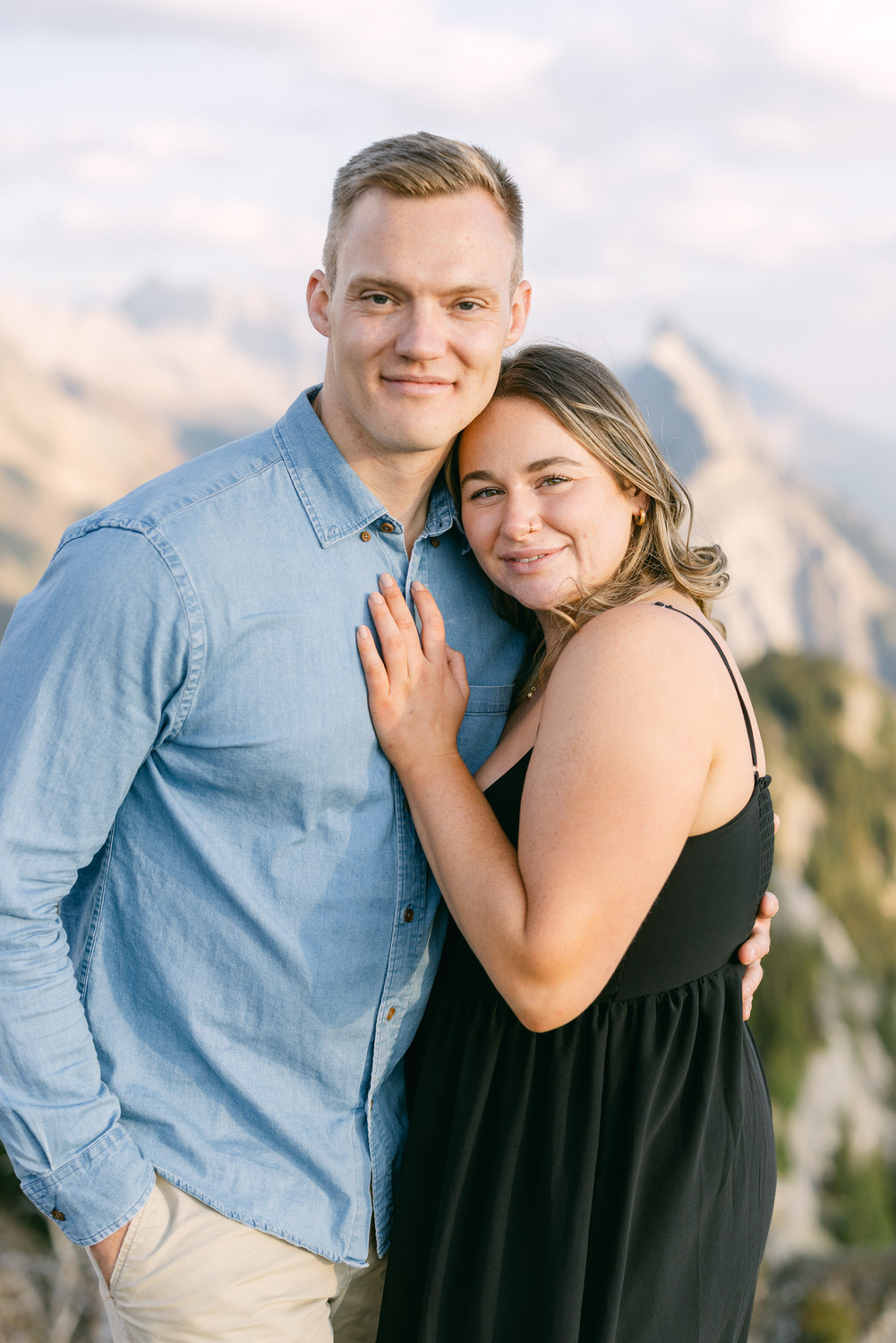 Couple Embracing in Nature::A young couple stands together in an embrace against a scenic mountain backdrop, showcasing warmth and connection.