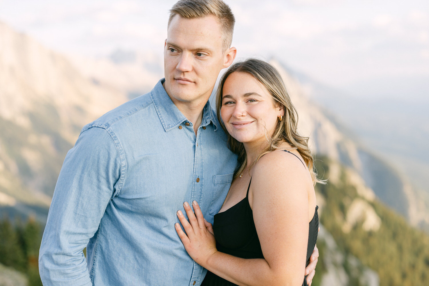 A happy couple poses together, with the woman smiling and resting her hand on the man's chest, against a backdrop of mountains and a bright sky.