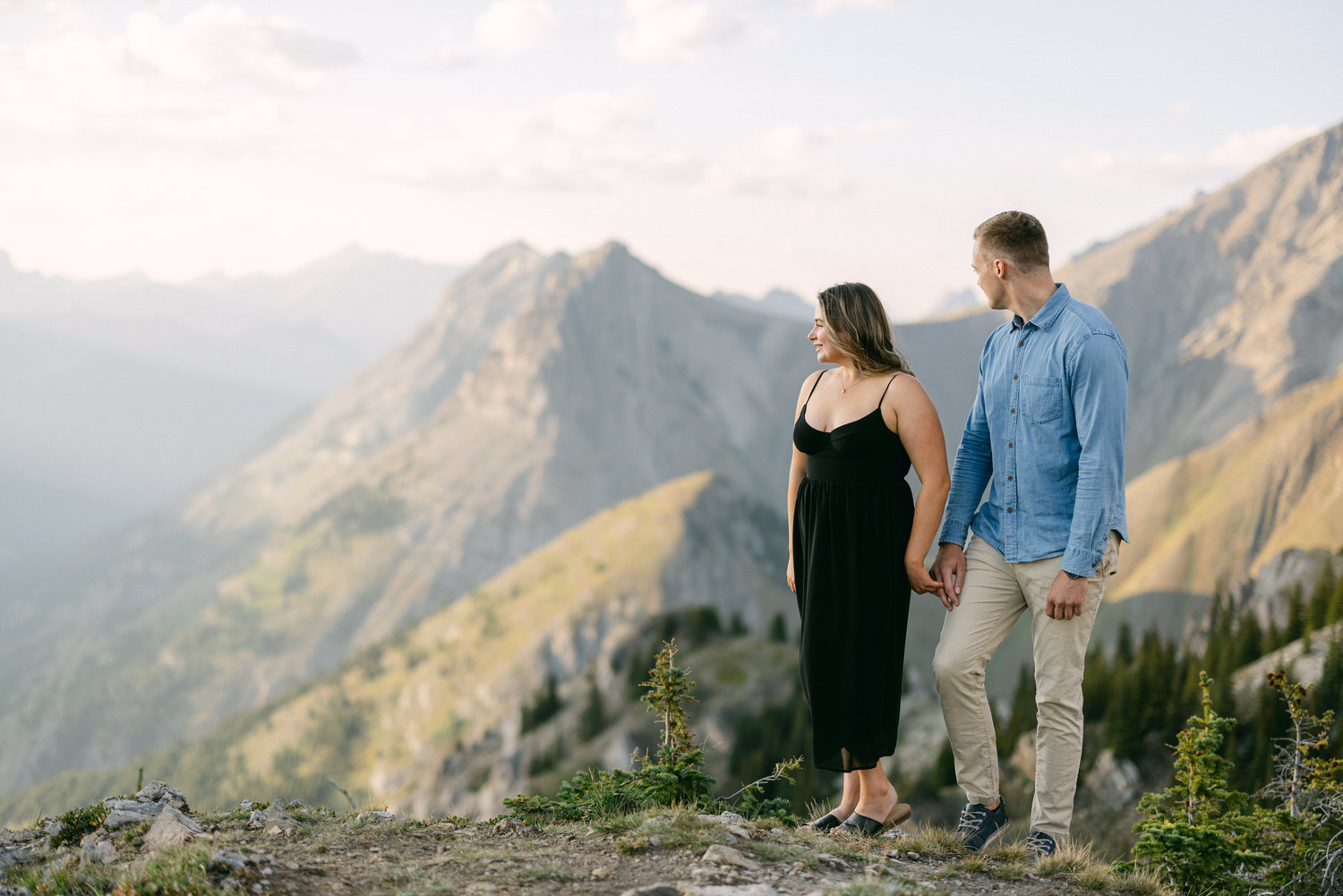 A couple holds hands while standing on a mountain overlook, gazing at the breathtaking scenery.