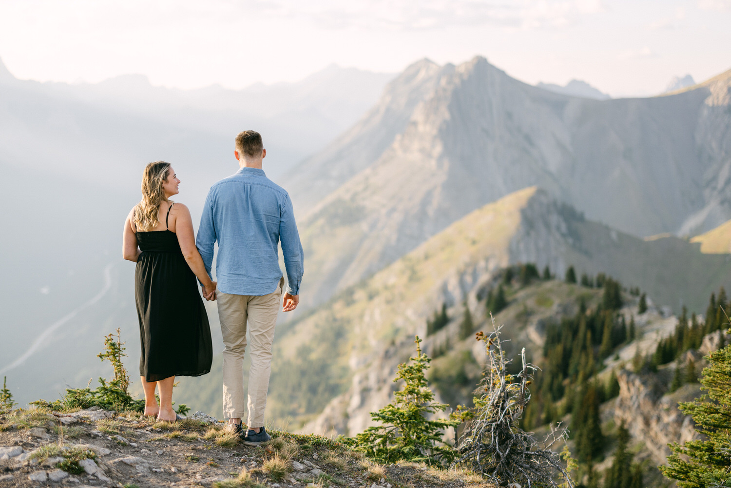 A couple holding hands while standing on a mountain peak, surrounded by dramatic landscapes and soft sunlight.