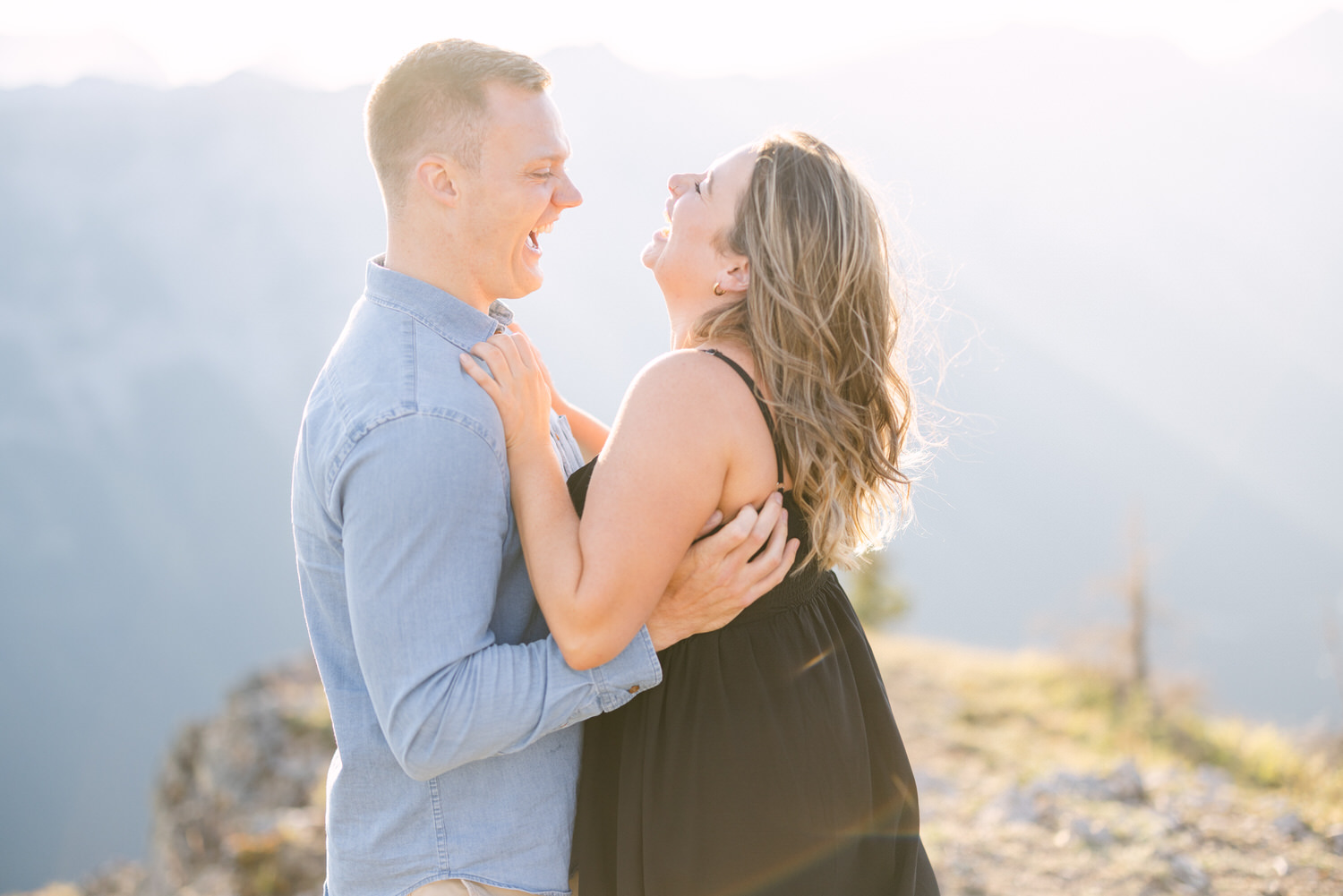 A happy couple laughs and embraces outdoors with a scenic mountain backdrop during golden hour.