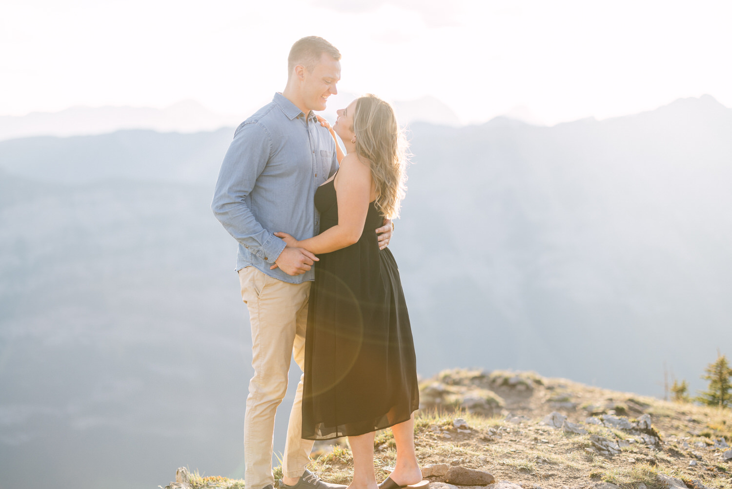 A couple shares a tender moment on a mountaintop, surrounded by a breathtaking view, with soft sunlight illuminating their connection.