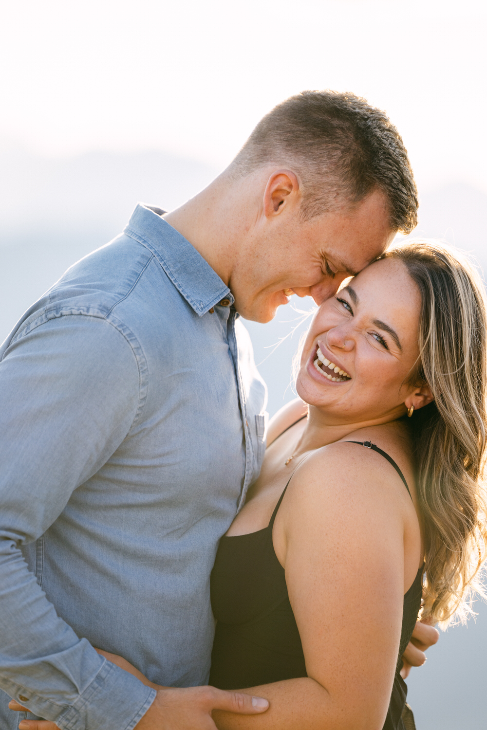 A couple shares a joyful moment outdoors, laughing and embracing each other, with a soft, sunlit backdrop creating a warm atmosphere.