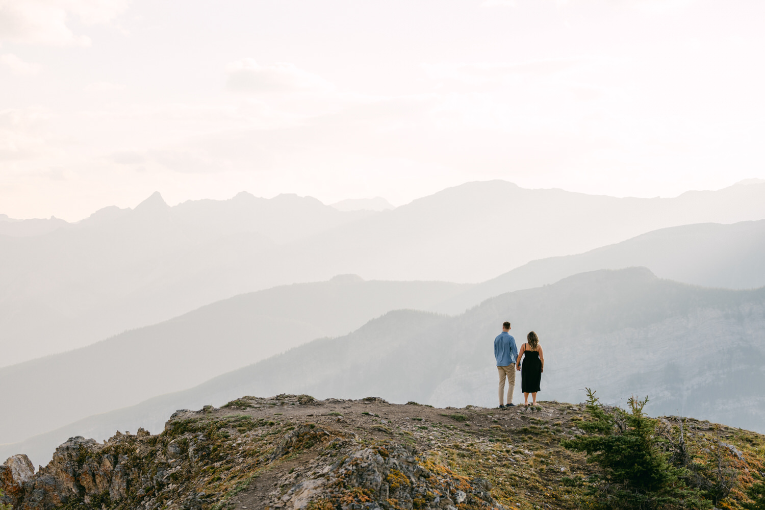 Serene Mountain Escape::A couple stands hand in hand on a rocky ledge overlooking misty mountains in the distance during golden hour.