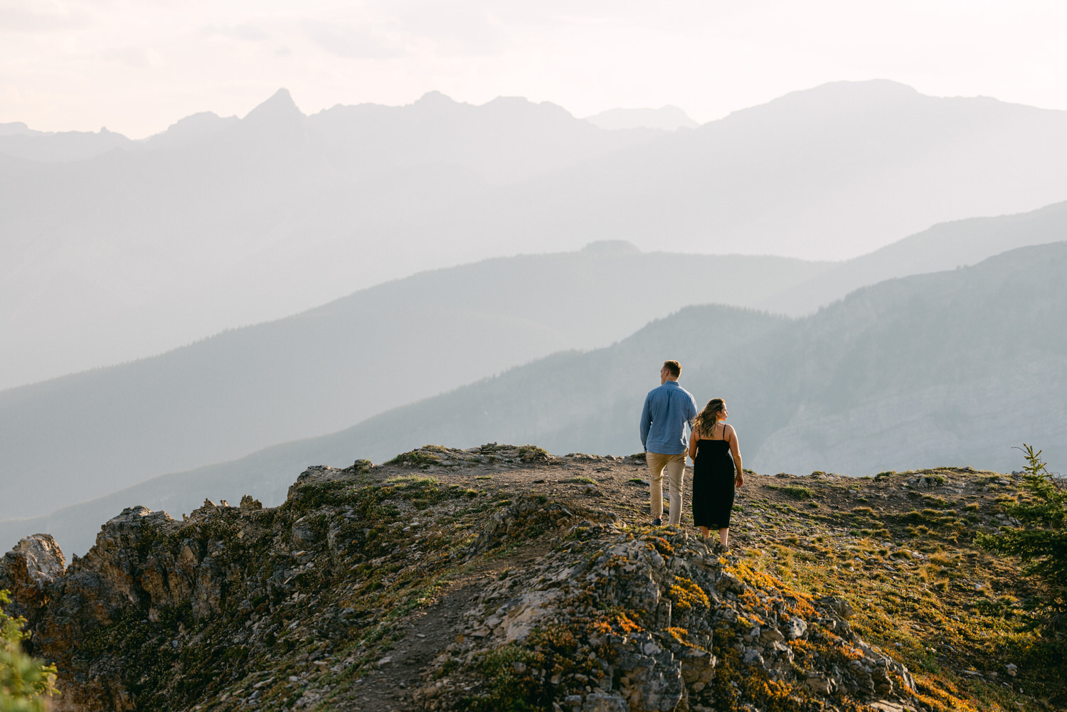 A couple stands hand-in-hand atop a rugged mountain ridge, gazing at the distant misty peaks and valleys during golden hour.