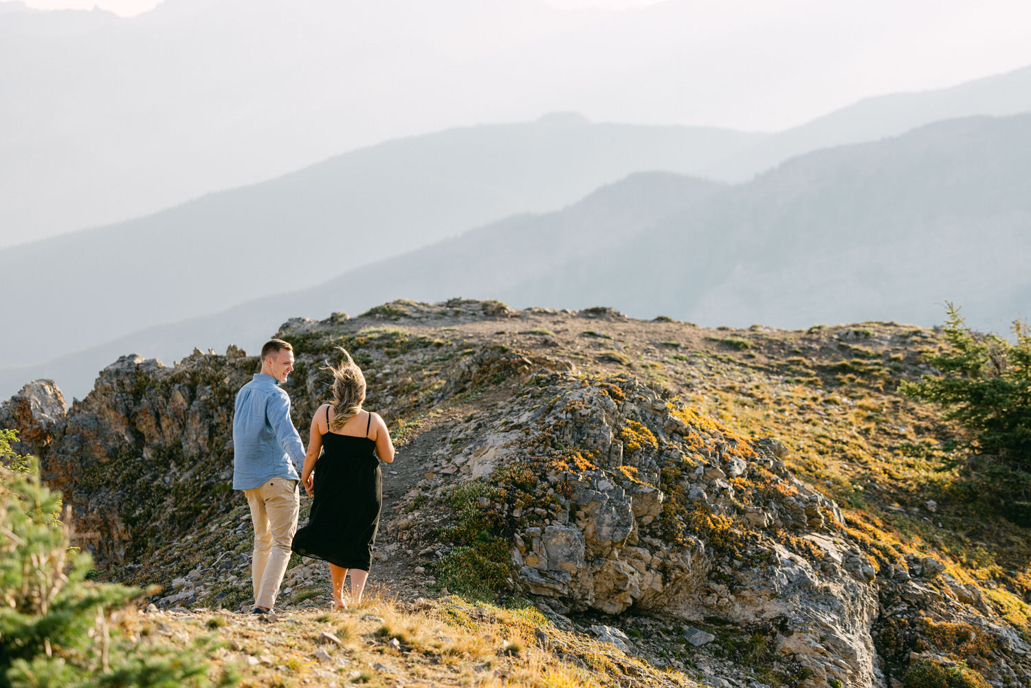 A couple walking hand in hand along a rocky mountain path with scenic mountains in the background.