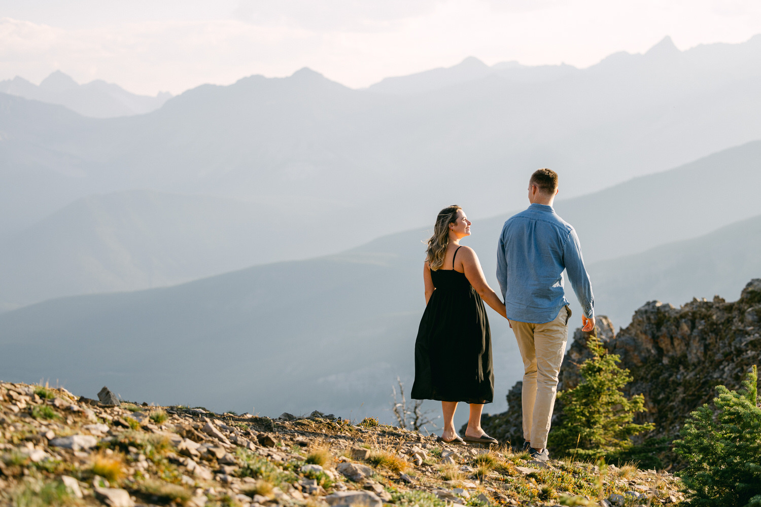 A couple walks hand in hand on a rocky path with a scenic mountain backdrop, bathed in soft sunlight.