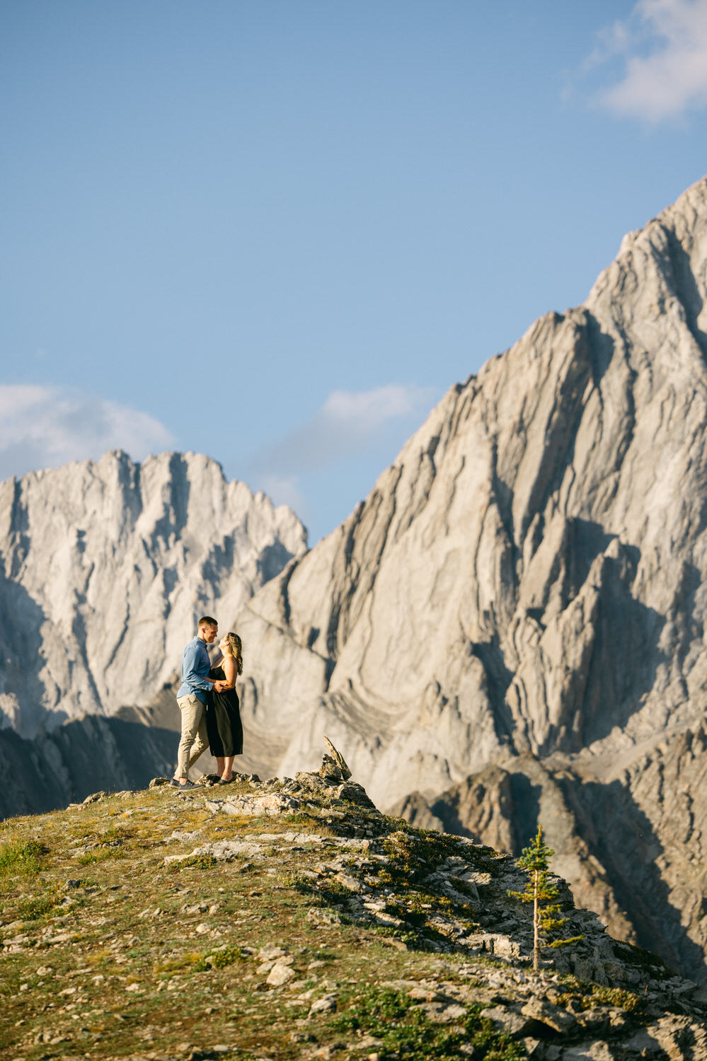 A couple stands together on a rocky outcrop surrounded by towering mountain peaks, basking in the warm glow of sunlight.