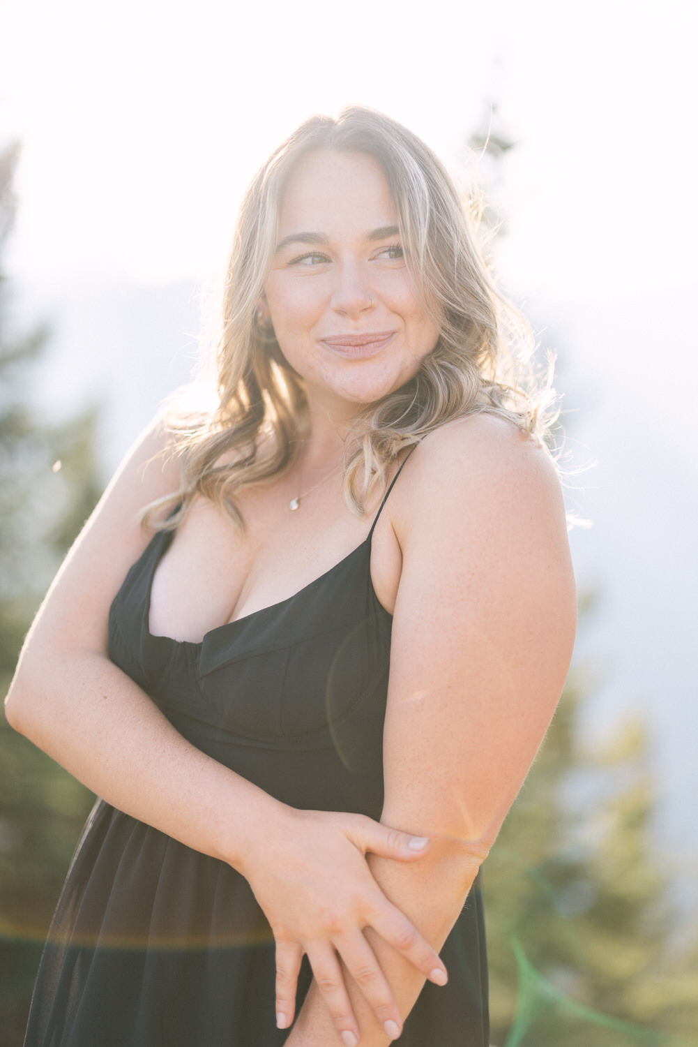 A smiling woman with long blonde hair poses gracefully outdoors, wearing a black dress against a bright, sunlit backdrop of trees and mountains.