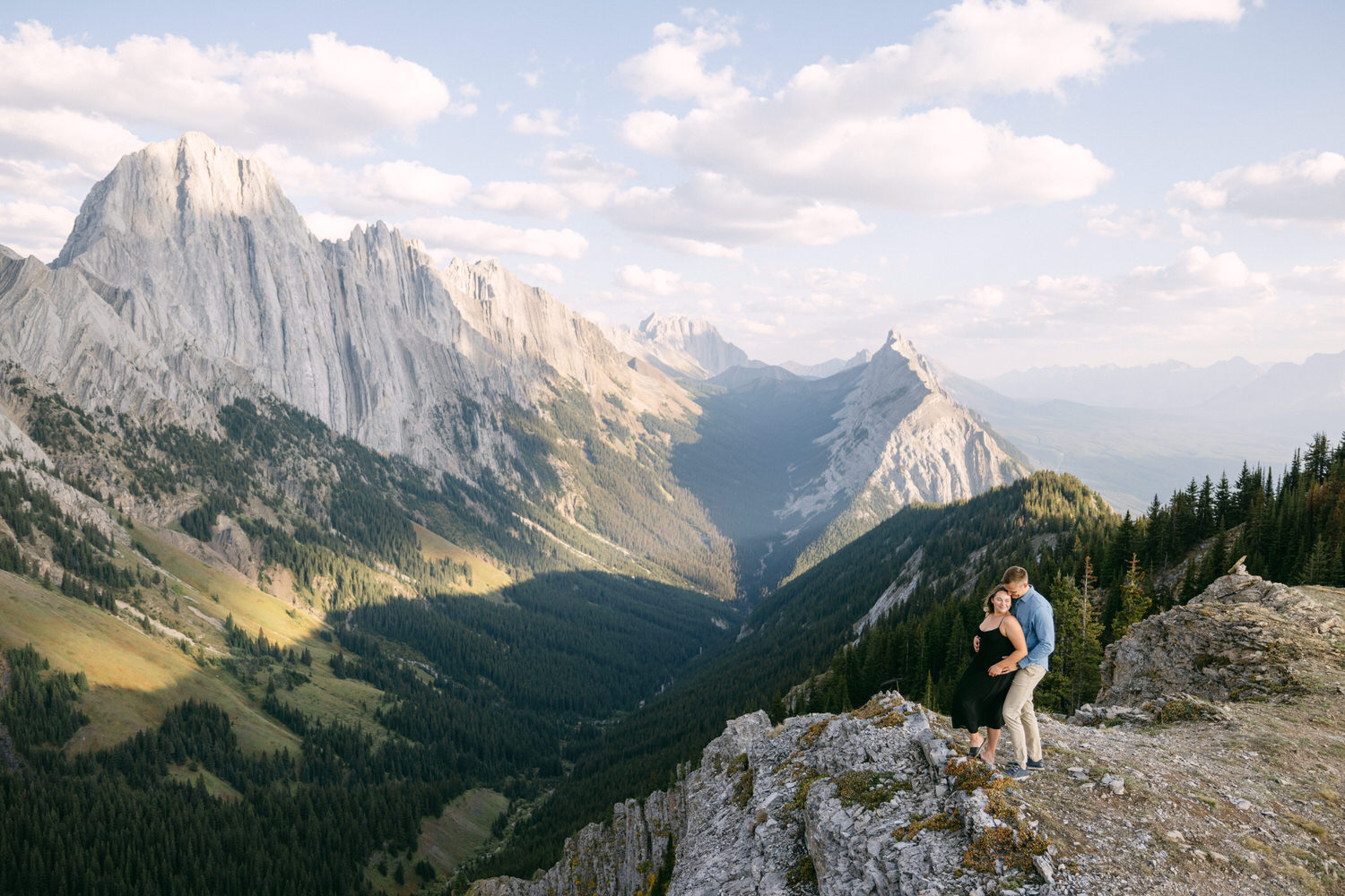 A couple embraces on a rocky outcrop with a breathtaking mountain backdrop under a partly cloudy sky.