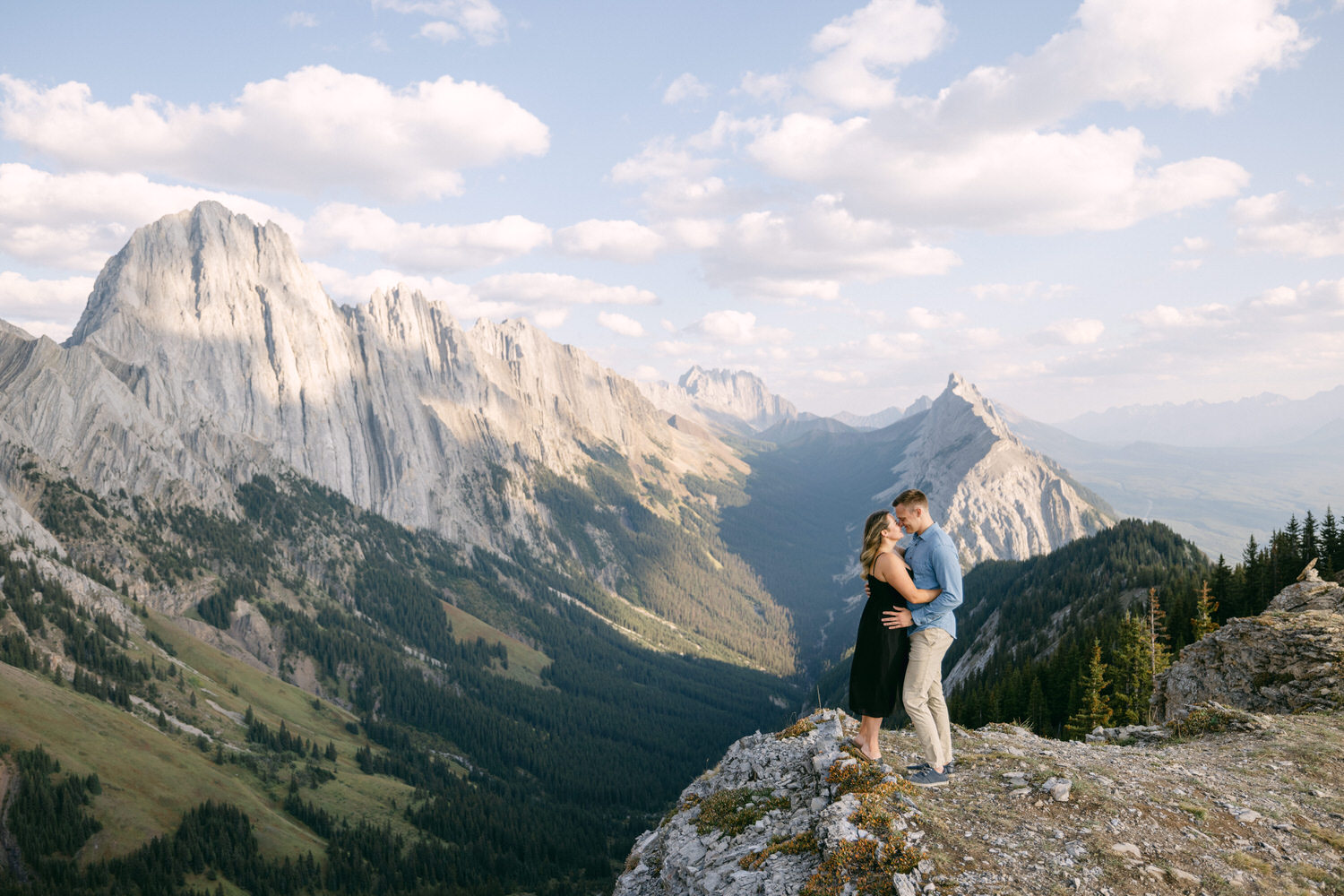 A couple embraces on a cliff with a dramatic mountain backdrop under a partly cloudy sky.