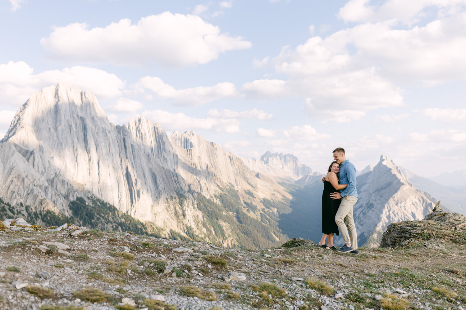 A couple hugs on a rocky outcrop with towering mountains in the background under a blue sky with clouds.