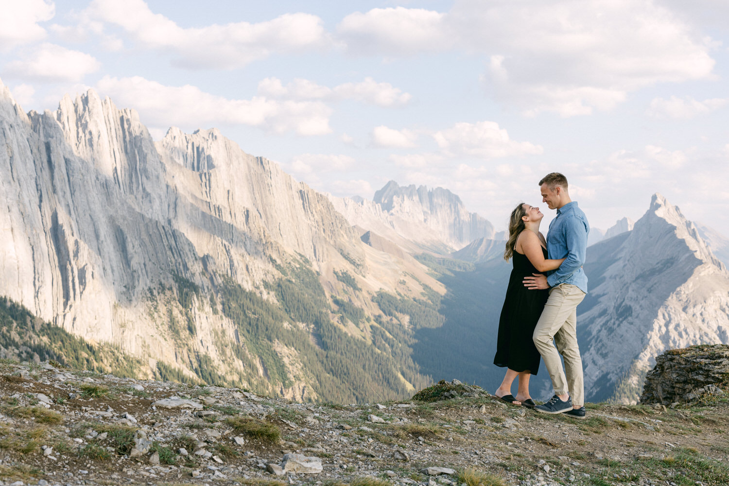 A couple joyfully embracing on a rocky mountaintop, surrounded by stunning mountain scenery and a bright sky.