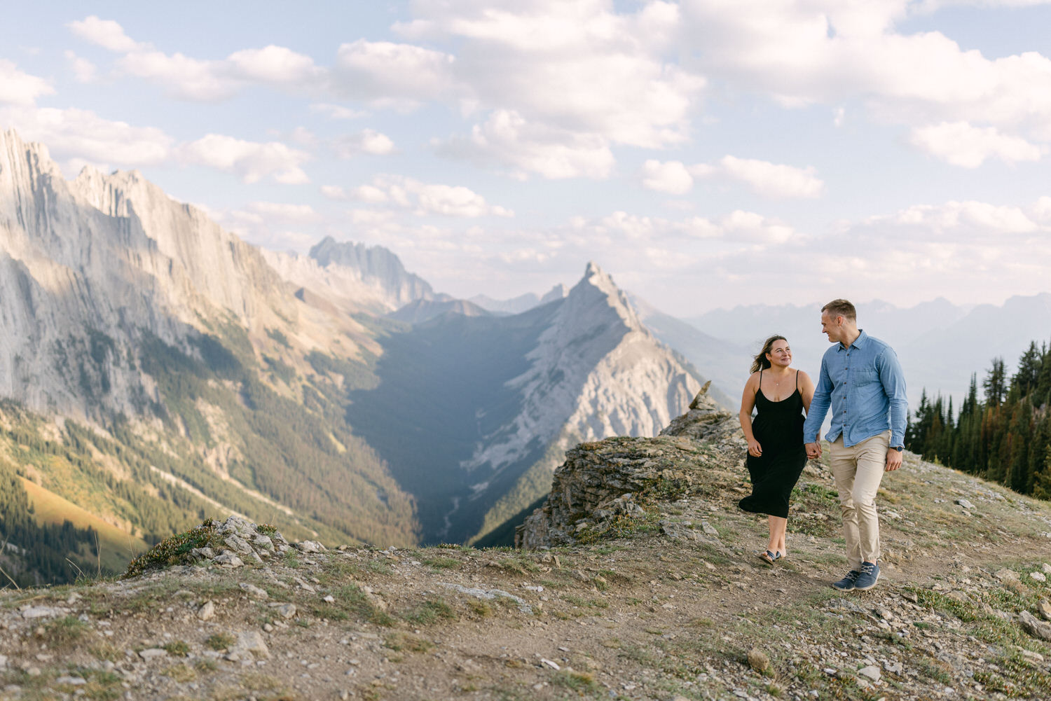 A man and woman stroll hand in hand along a rugged mountain path with dramatic peaks and lush greenery in the background.