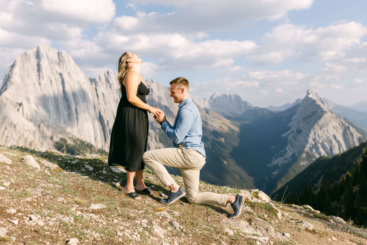 A joyous couple celebrates an engagement amidst breathtaking mountain scenery, with one partner kneeling and presenting a ring.