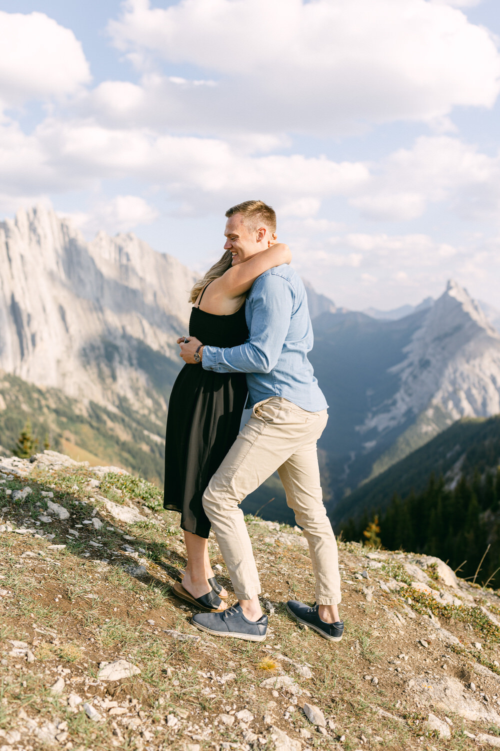 A happy couple sharing a warm embrace while enjoying breathtaking mountain views. The woman is wearing a black dress, and the man is in a blue shirt and beige pants, smiling against a stunning natural backdrop.