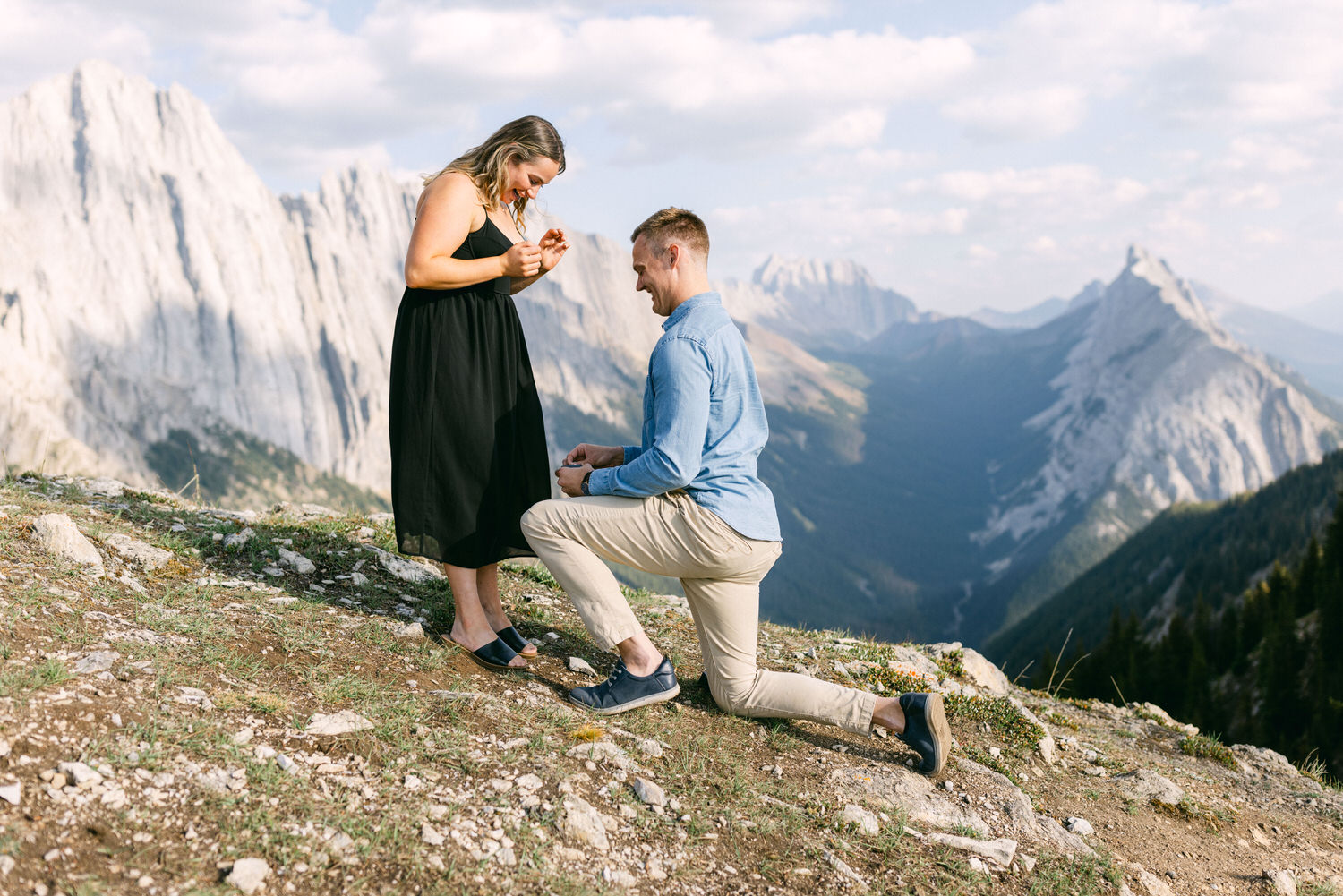 A man proposes to a woman while kneeling on a mountainside, with breathtaking peaks and a valley in the background.