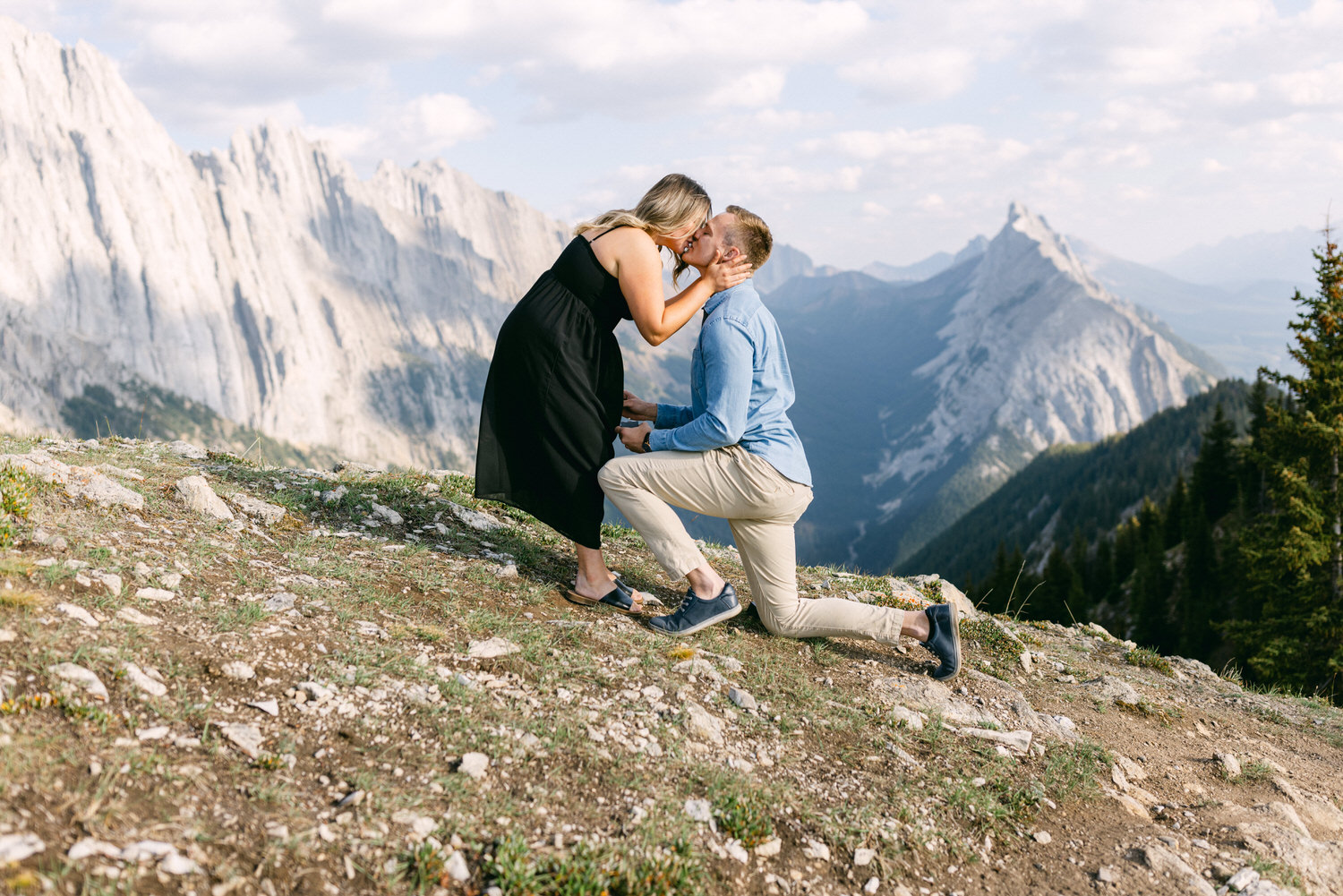 A man kneels to propose to a woman on a scenic mountainside, capturing a tender moment with breathtaking mountainous background.