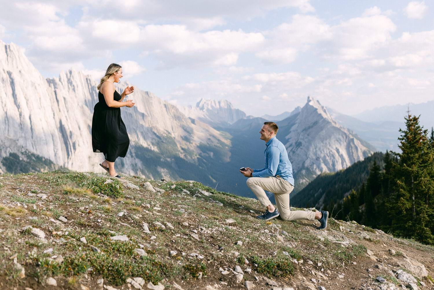 A man kneels with a ring, proposing to a woman who joyfully reacts in a scenic mountainous landscape.