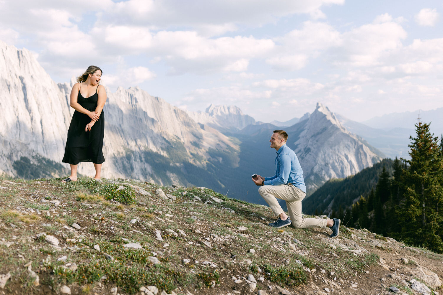 A joyful couple during a mountain proposal, with the man on one knee and the woman smiling in a stunning mountainous backdrop.