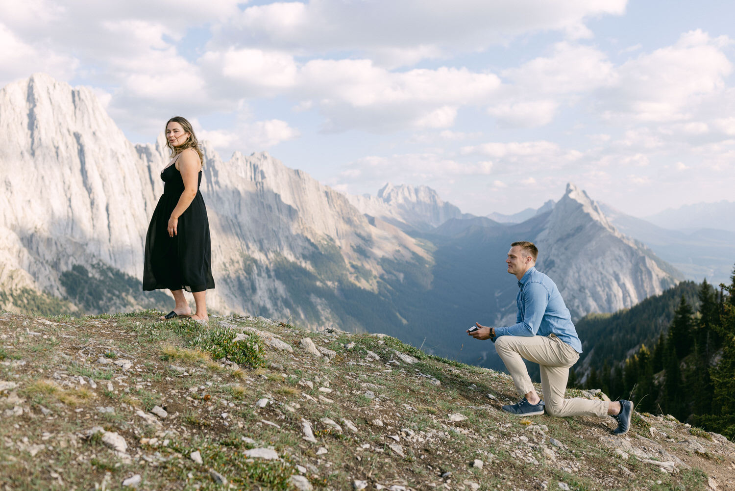 A woman stands gracefully on a hillside, while a man kneels beside her, holding a ring, against a majestic mountain backdrop.