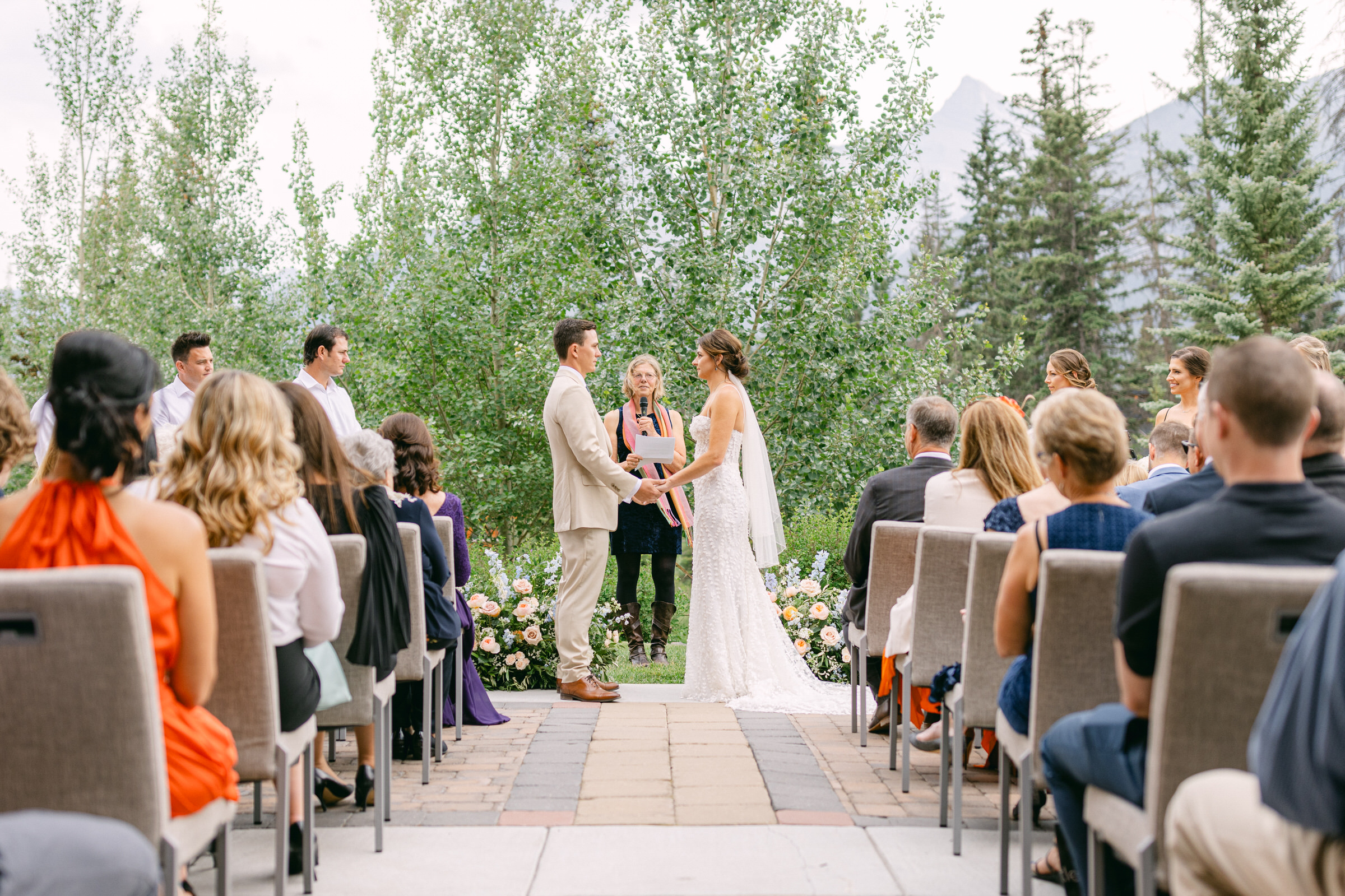 A couple exchanges vows during an outdoor wedding ceremony surrounded by guests and lush greenery.