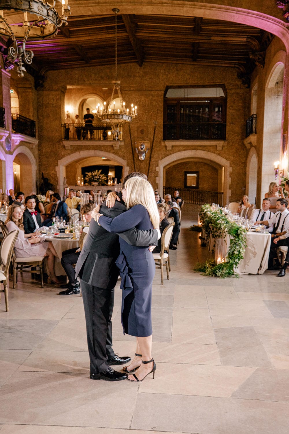 A man and a woman embrace during a dance at a formal event, surrounded by elegantly dressed guests seated at tables in a beautifully decorated venue.