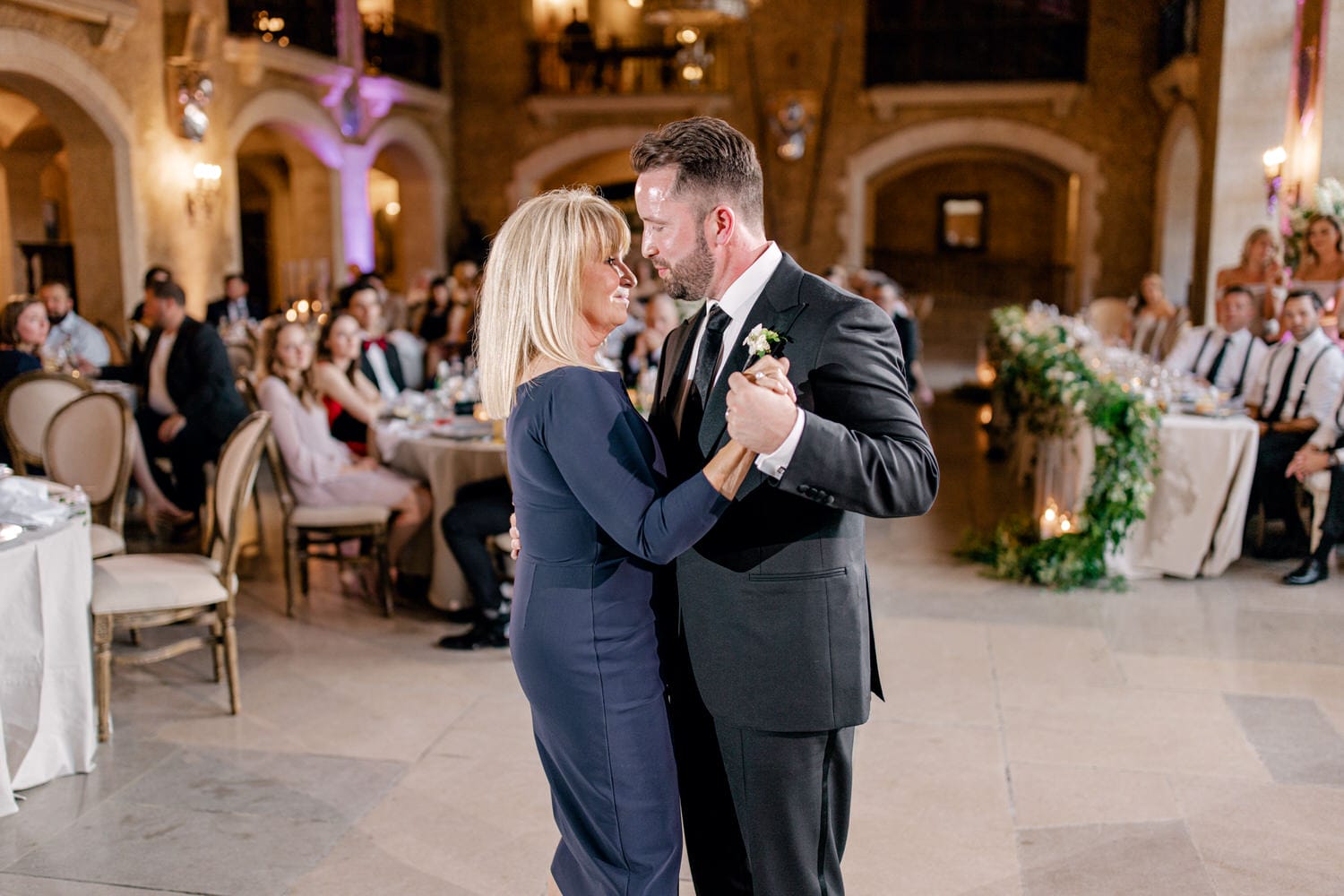 A touching moment between a mother and son as they dance together at a wedding reception, surrounded by guests enjoying the celebration.