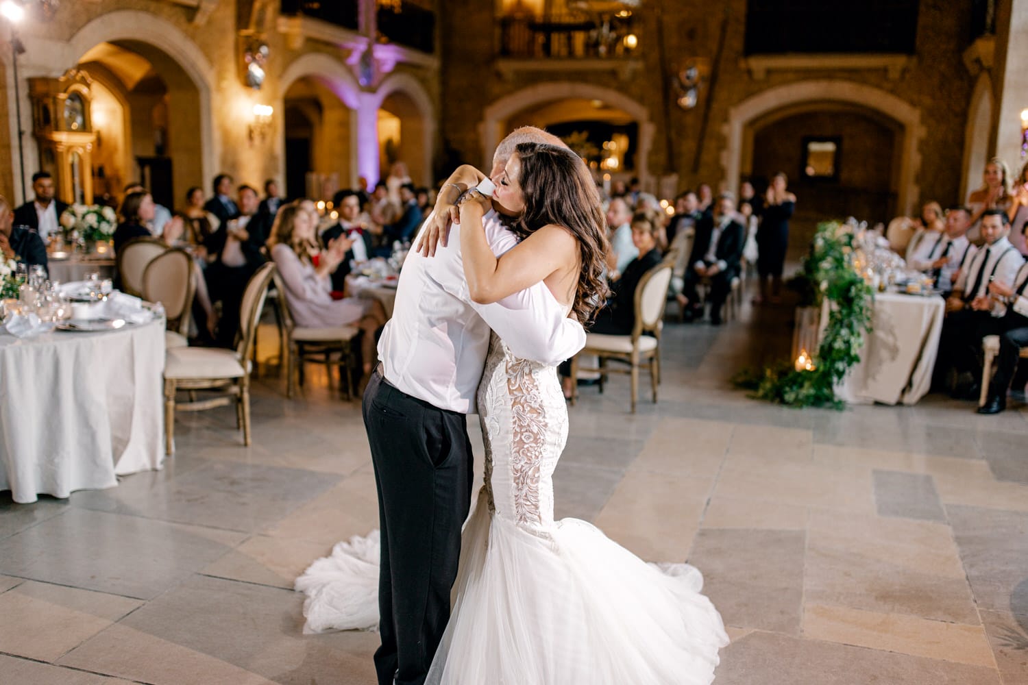 A newly married couple embraces during their first dance in a beautifully decorated reception hall, surrounded by guests clapping and celebrating.