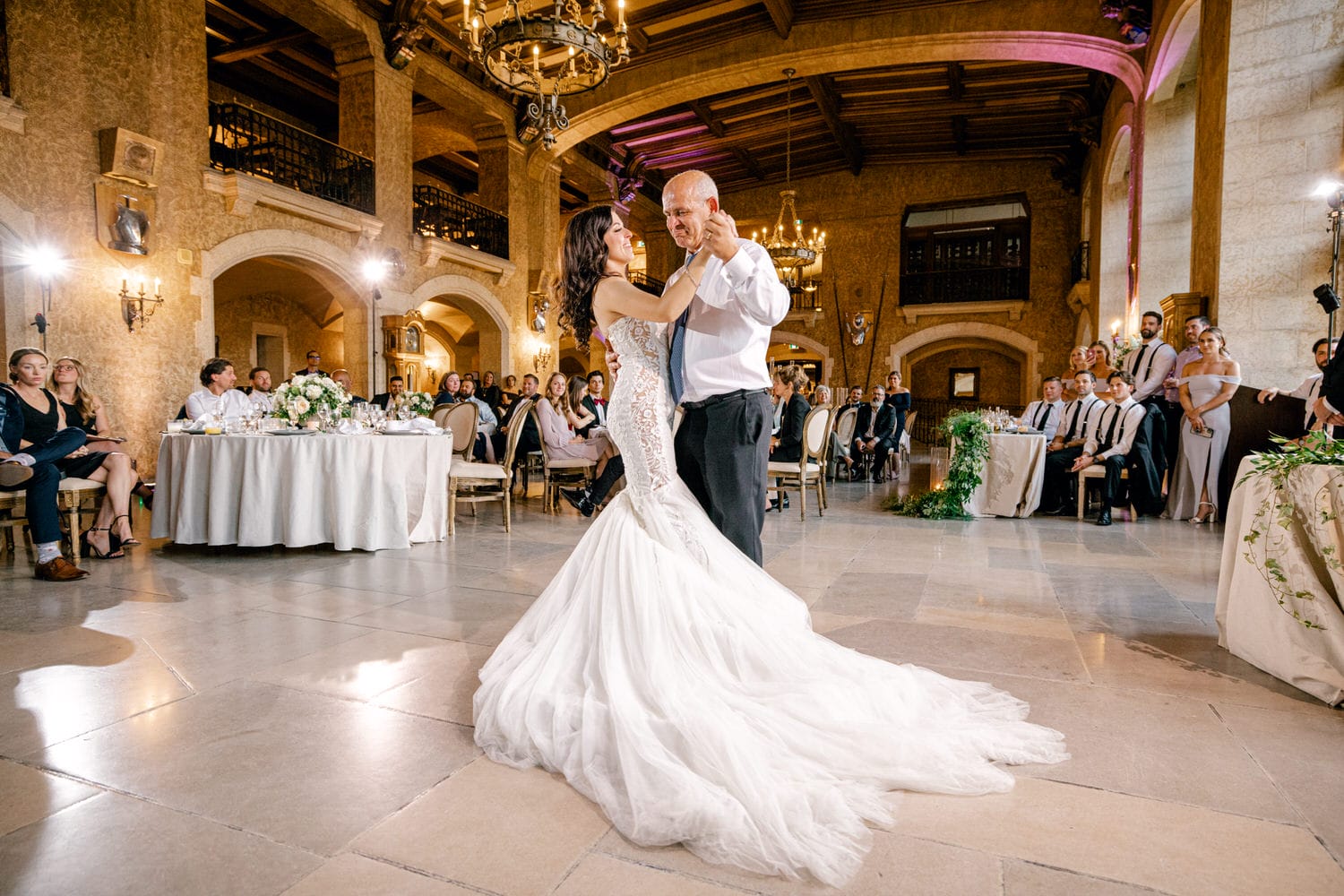 A bride and her father share a heartfelt dance during a wedding reception, surrounded by guests in an elegantly decorated venue.