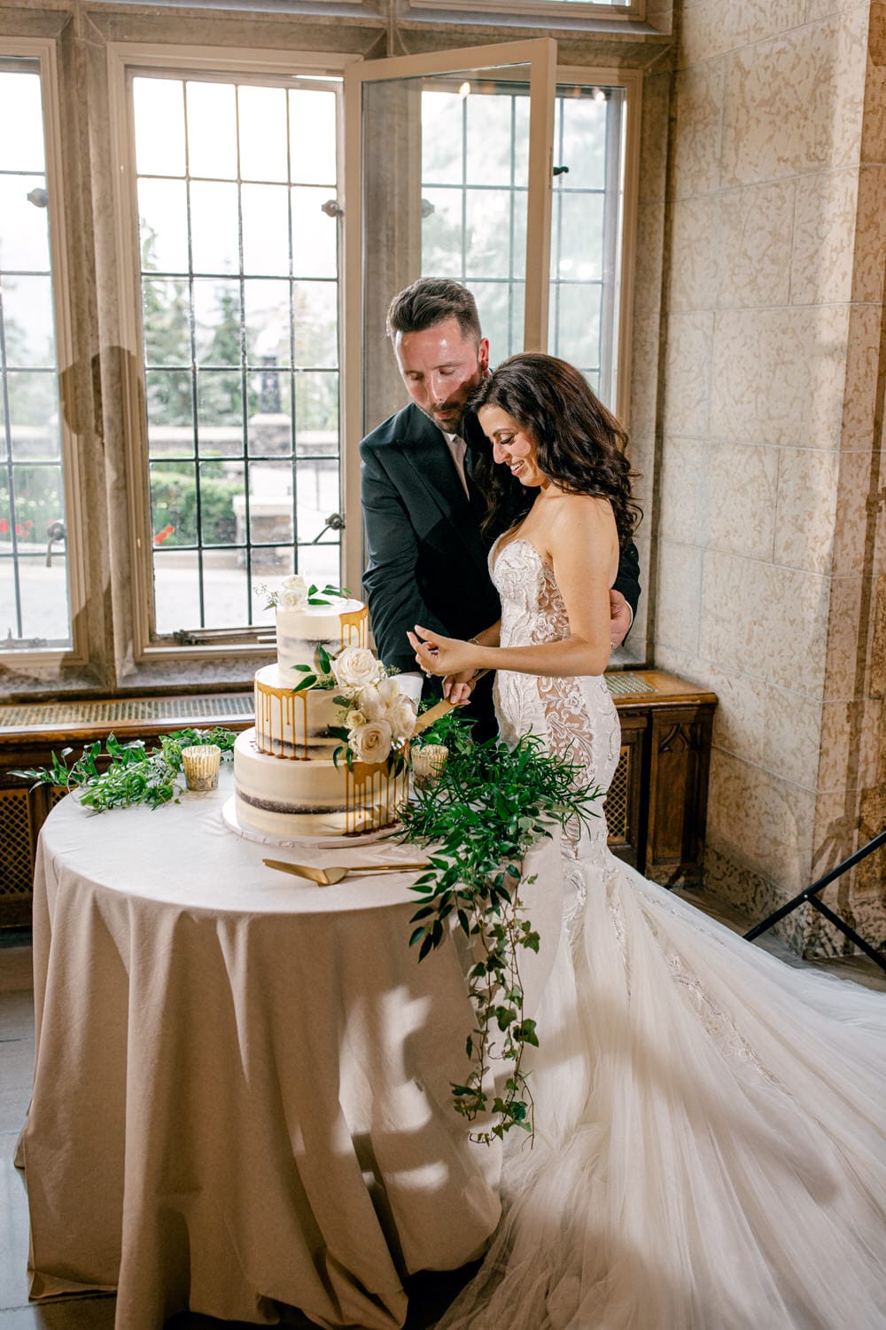 A couple joyfully slicing their wedding cake adorned with flowers and greenery, set against a backdrop of large windows.