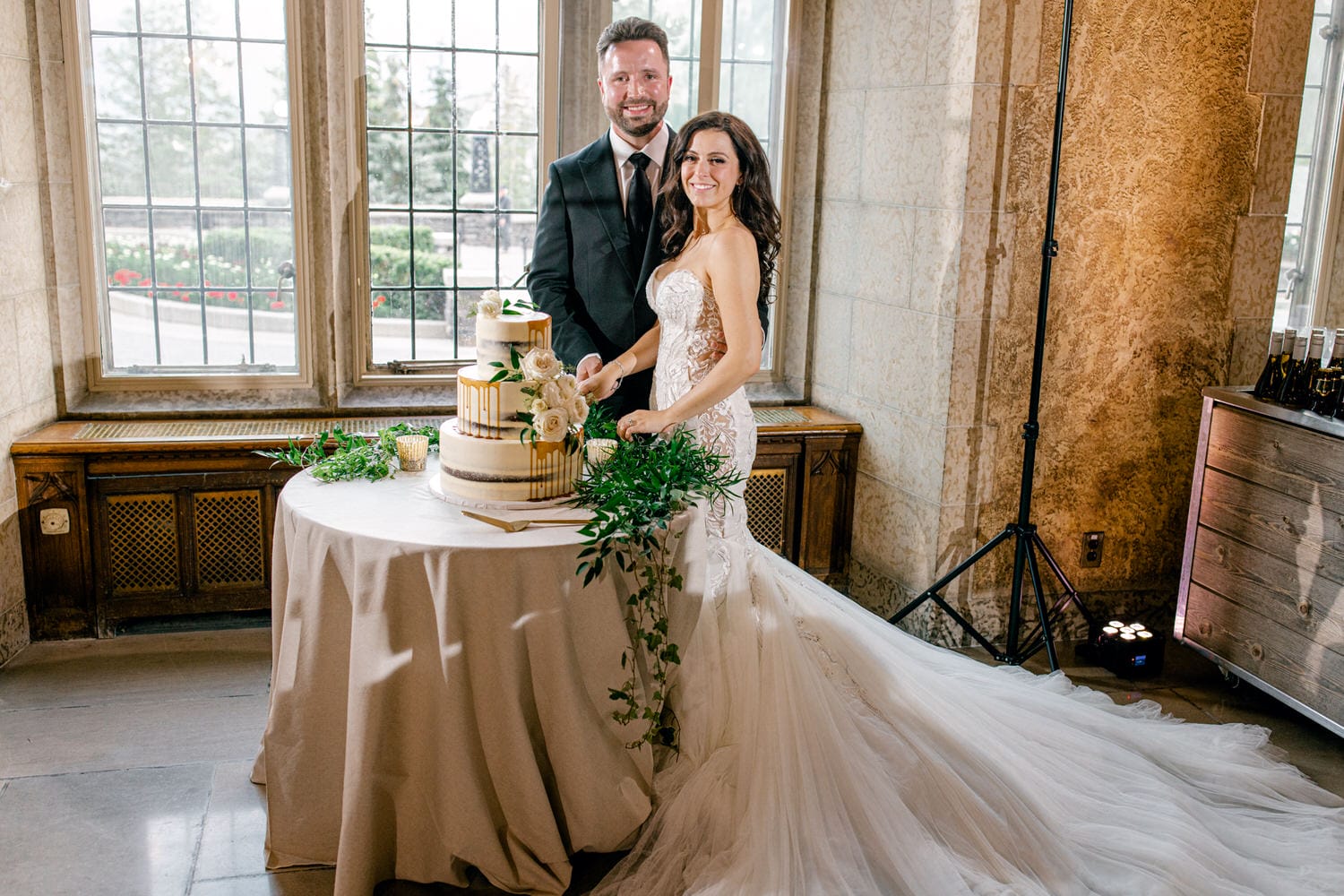 A couple smiles together as they prepare to cut their wedding cake, adorned with flowers, in an elegant venue with large windows and soft lighting.