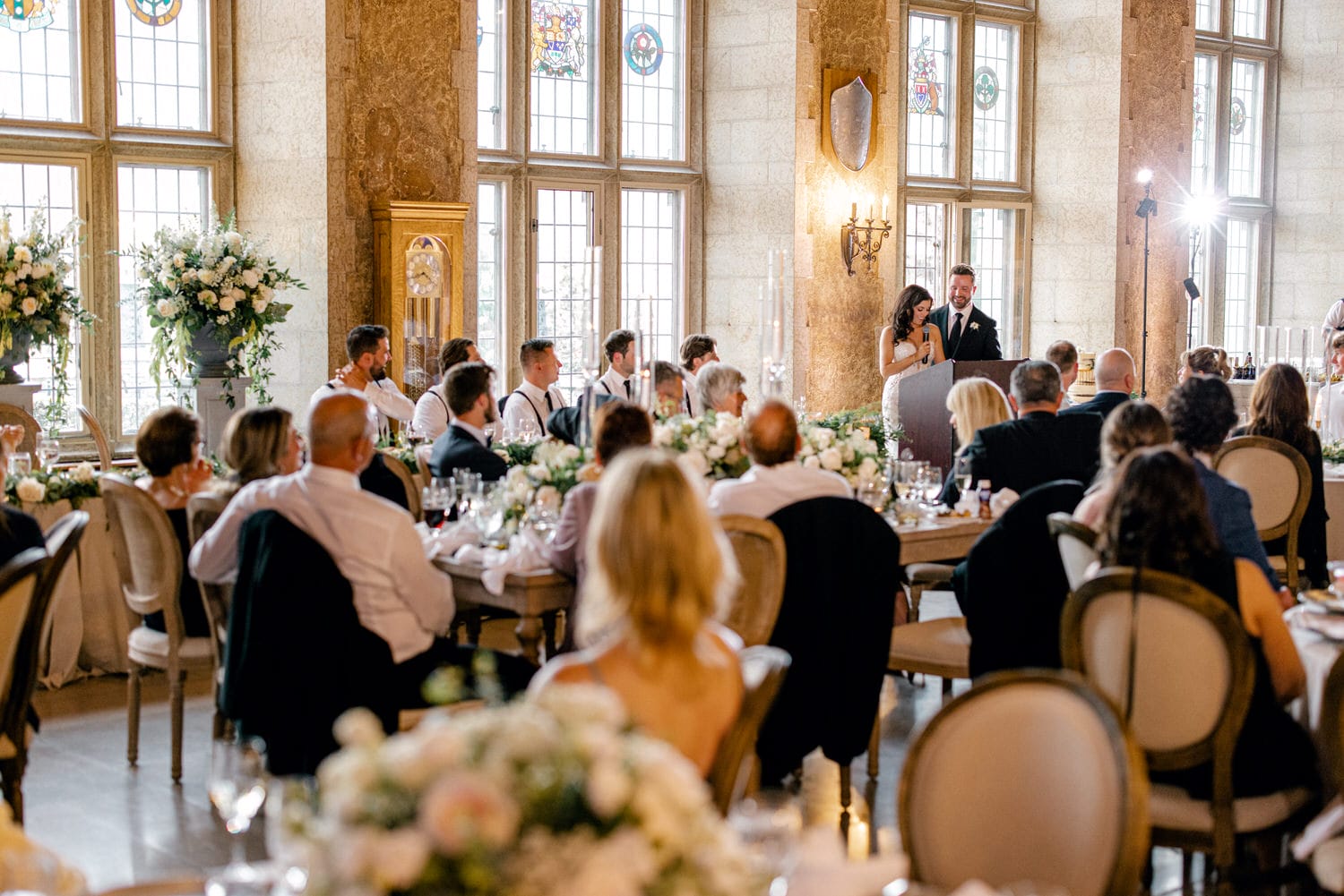 Guests enjoying heartfelt speeches at an elegant wedding reception, surrounded by floral decorations and stained glass windows.