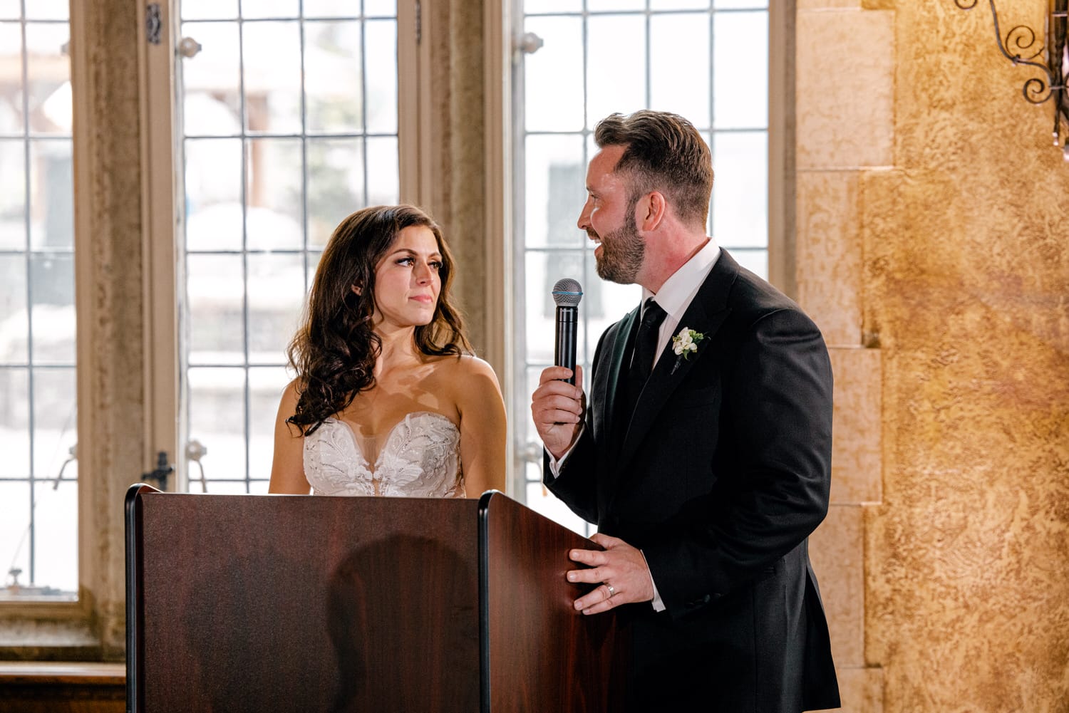 A bride and groom stand at a podium, sharing intimate smiles as the groom speaks into a microphone during their wedding reception. Elegant decor and large windows create a warm ambiance.