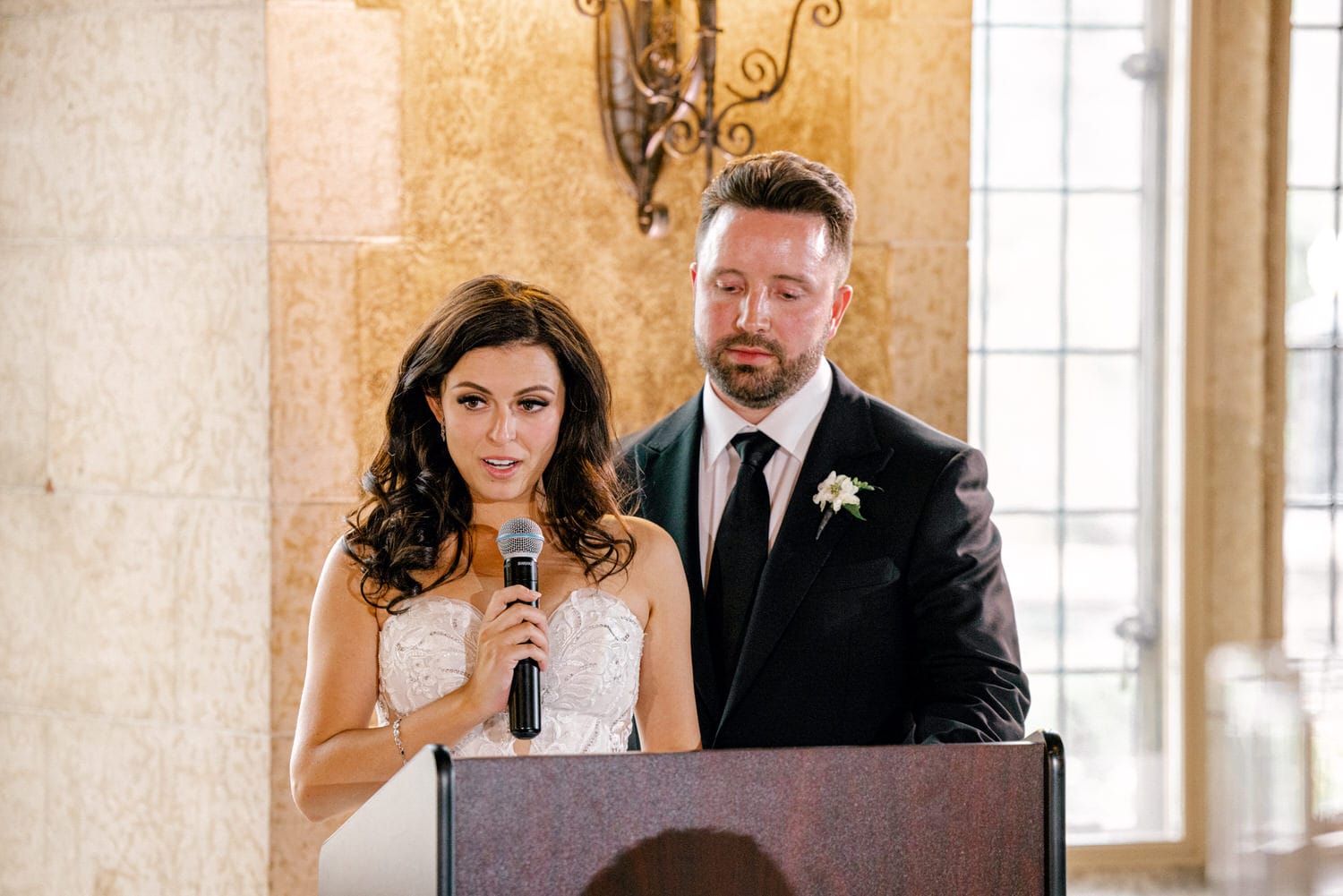 A bride speaks into a microphone while a suited man stands beside her, both in an elegantly decorated venue.