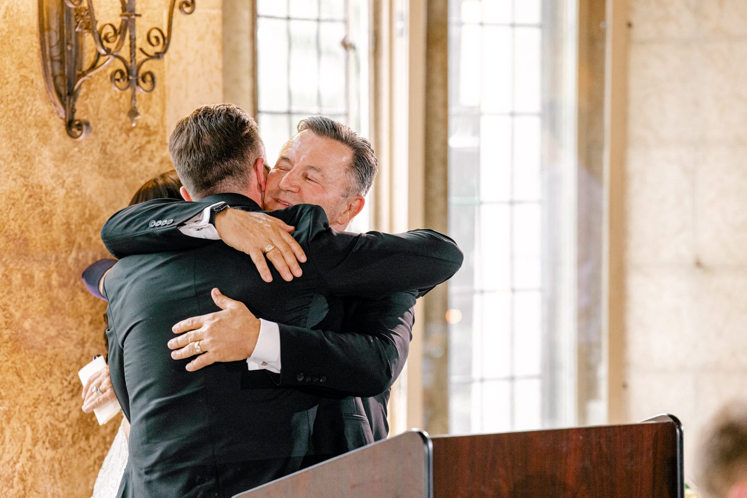 Two men in formal attire share a heartfelt hug, conveying warmth and camaraderie in a beautifully lit indoor setting.